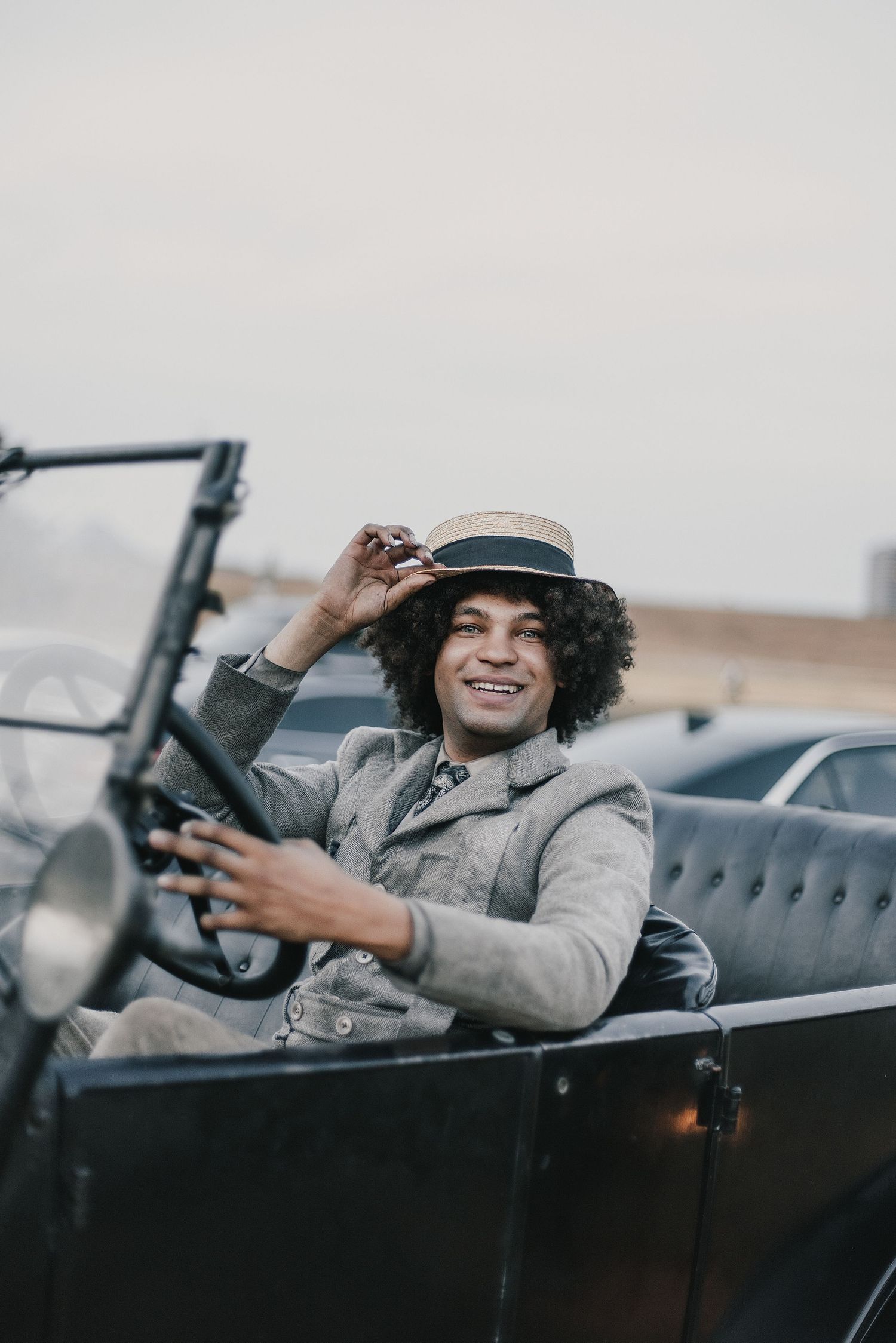 Smiling man with curly hair wearing a straw boater hat driving a vintage classic car outdoors.