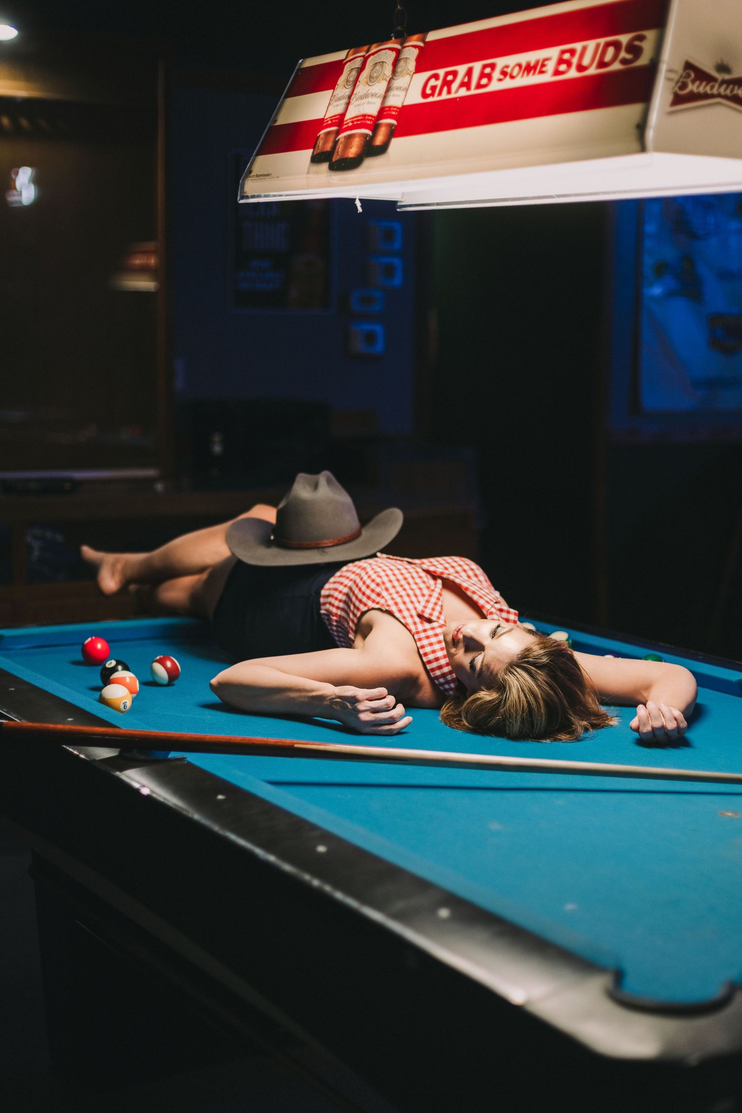 Woman in red shirt lying on a blue pool table under a Budweiser light in a dimly lit bar.