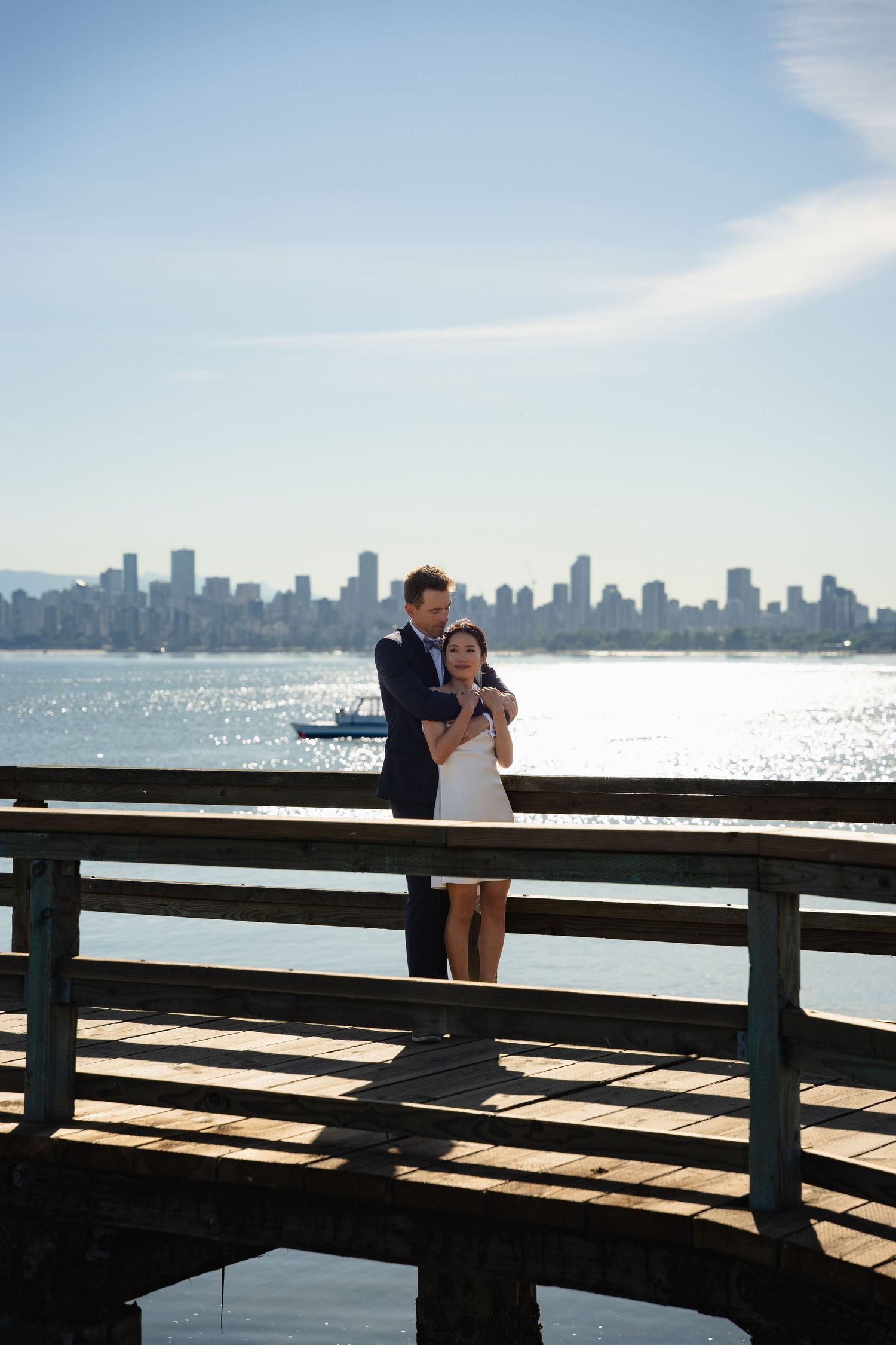 Couple embracing on wooden pier with Vancouver city skyline and blue water in background on sunny day.