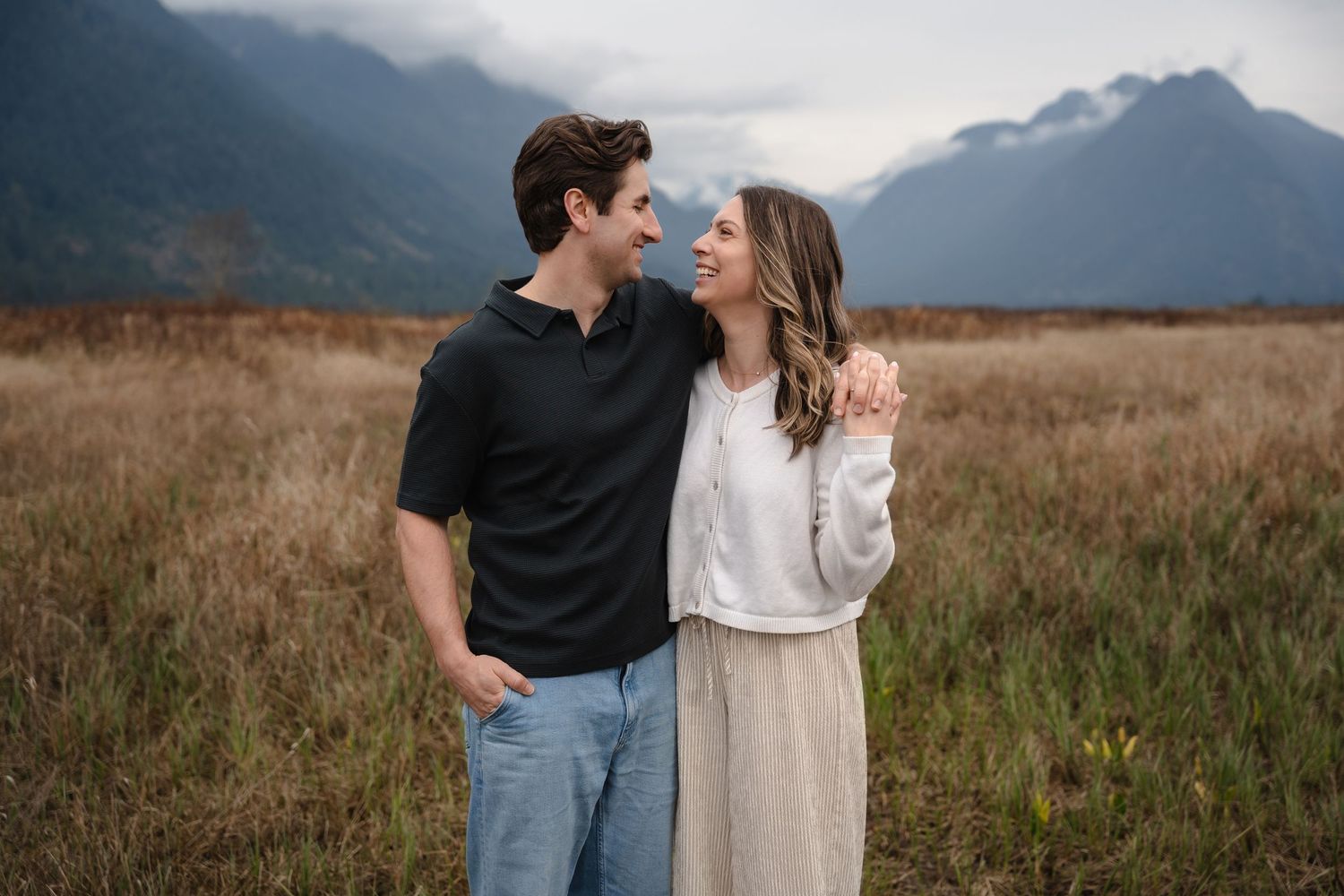 Couple smiling at each other in a golden field with misty mountains in the background.