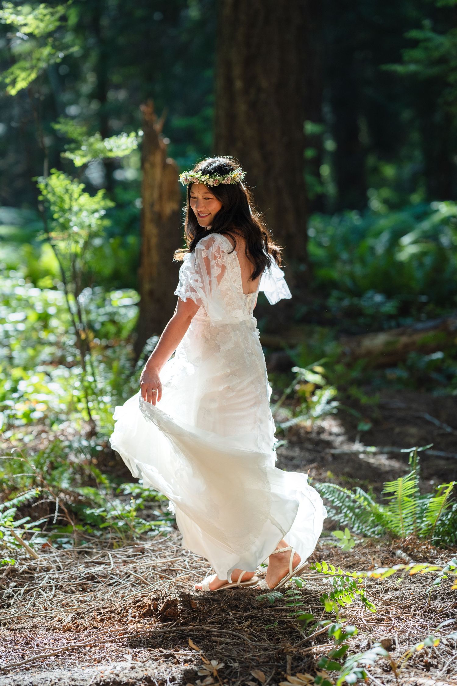 Smiling bride in white lace dress and floral crown twirling in a sunlit forest.