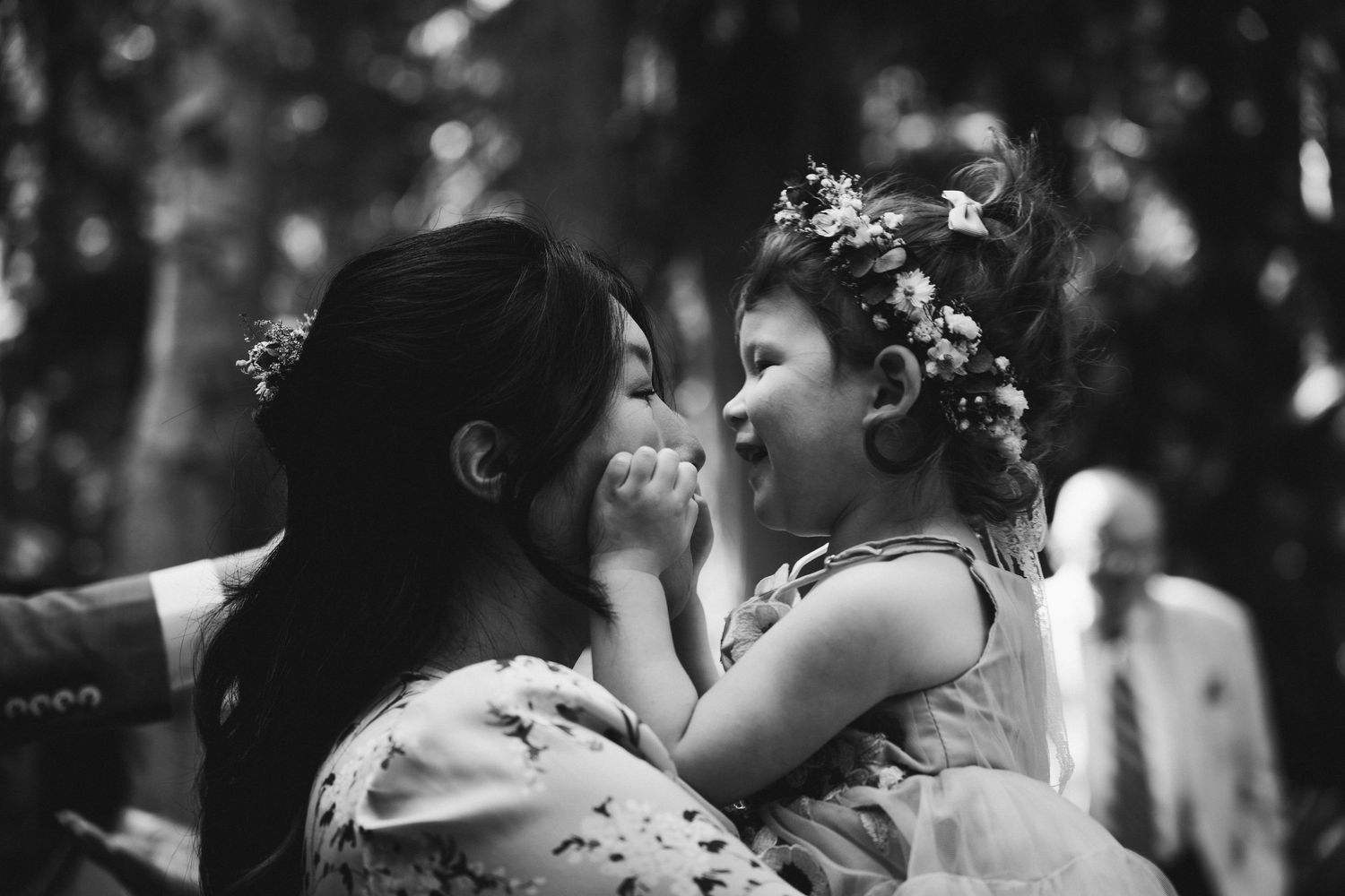 Black and white photo of a woman in floral dress holding a laughing little girl wearing a flower crown outdoors.