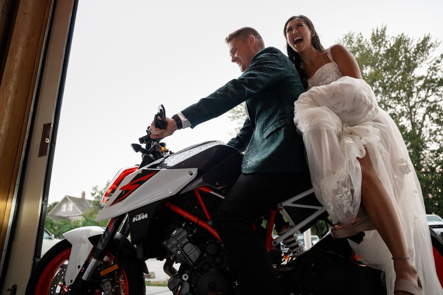 Bride in white dress and groom in green suit laughing together on a red and white KTM motorcycle.