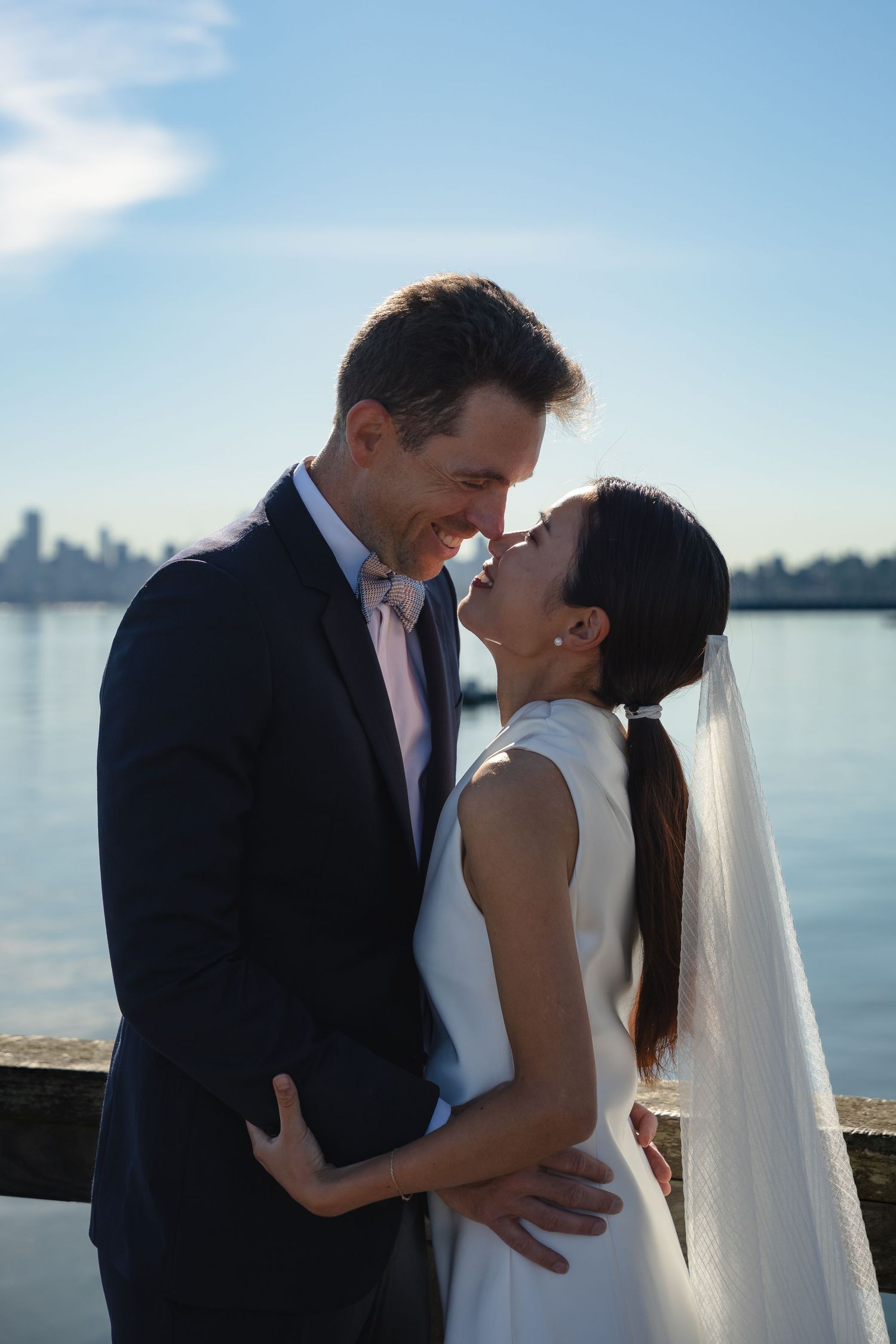Bride and groom share a romantic moment by the waterfront, with a city skyline in the background on a sunny day.