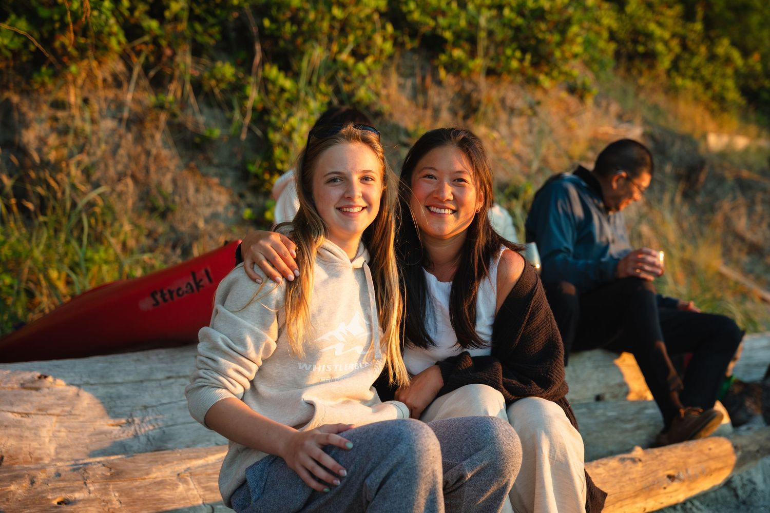 Two smiling young women sit together on a log at a sunny beach with a red canoe and friends in the background.