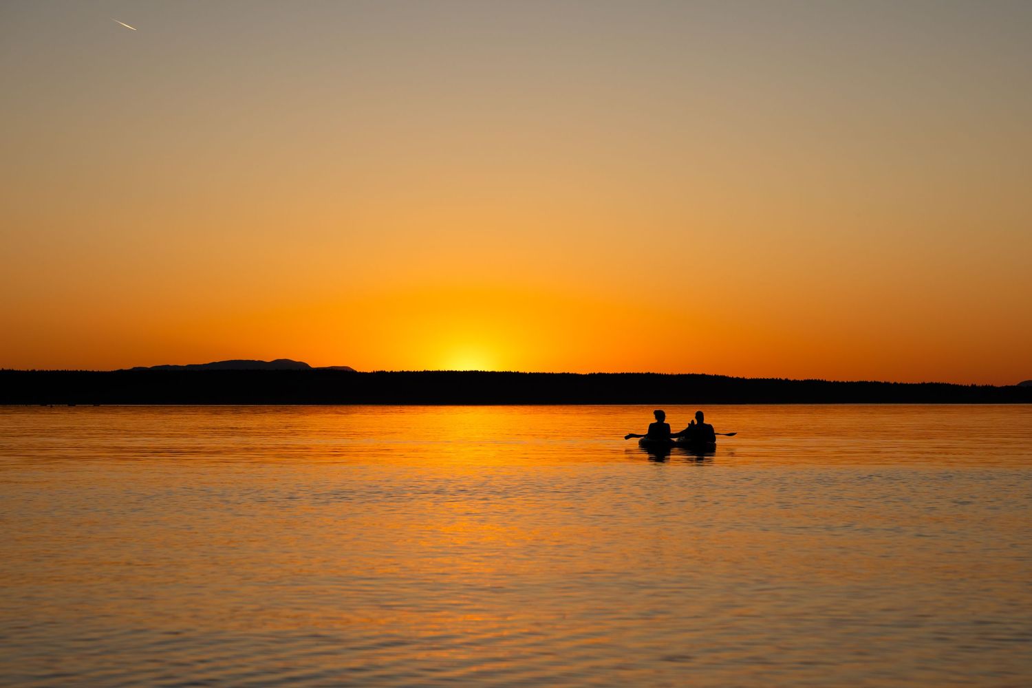 Two people kayaking on calm water silhouetted against a vibrant golden orange sunset over the horizon.