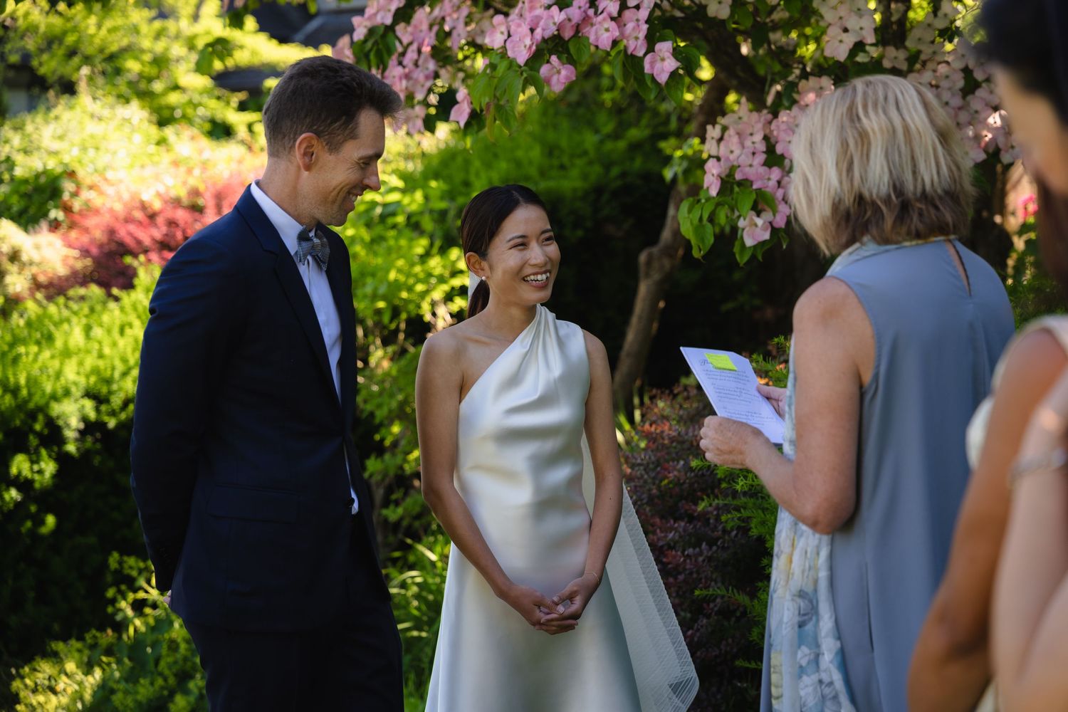 Couple exchanging vows outdoors in a garden wedding ceremony with pink blossoms and a female officiant.