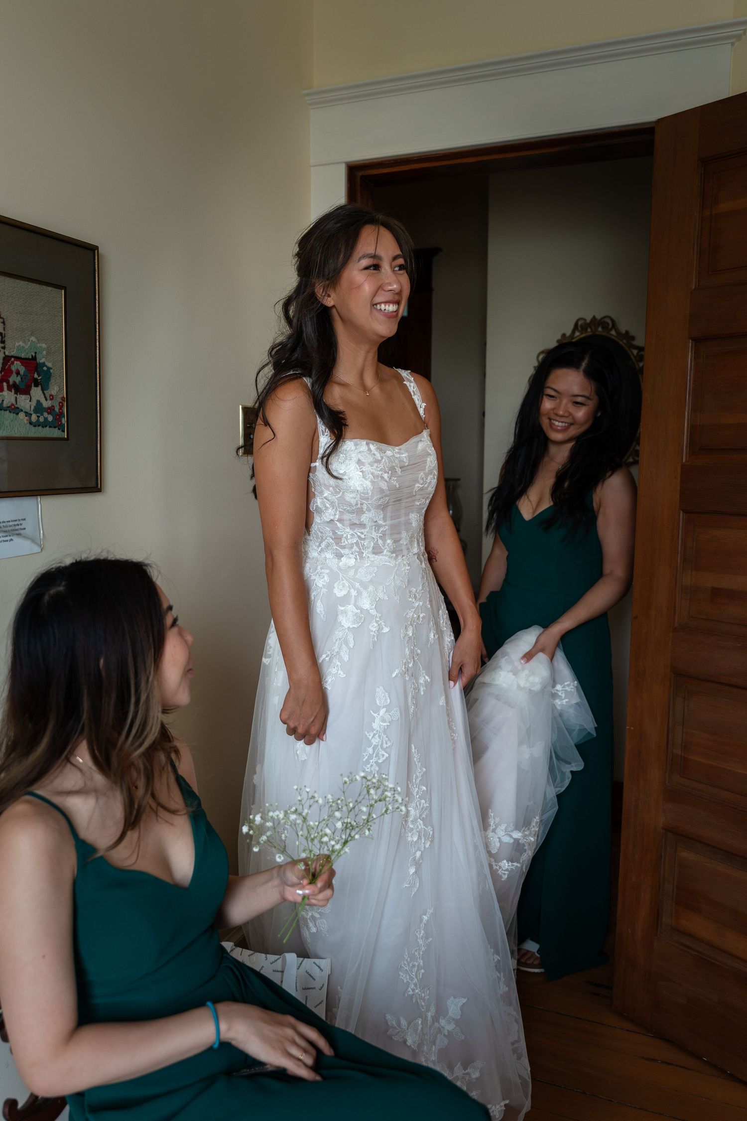 Smiling bride in white lace gown with bridesmaids in emerald green dresses getting ready.