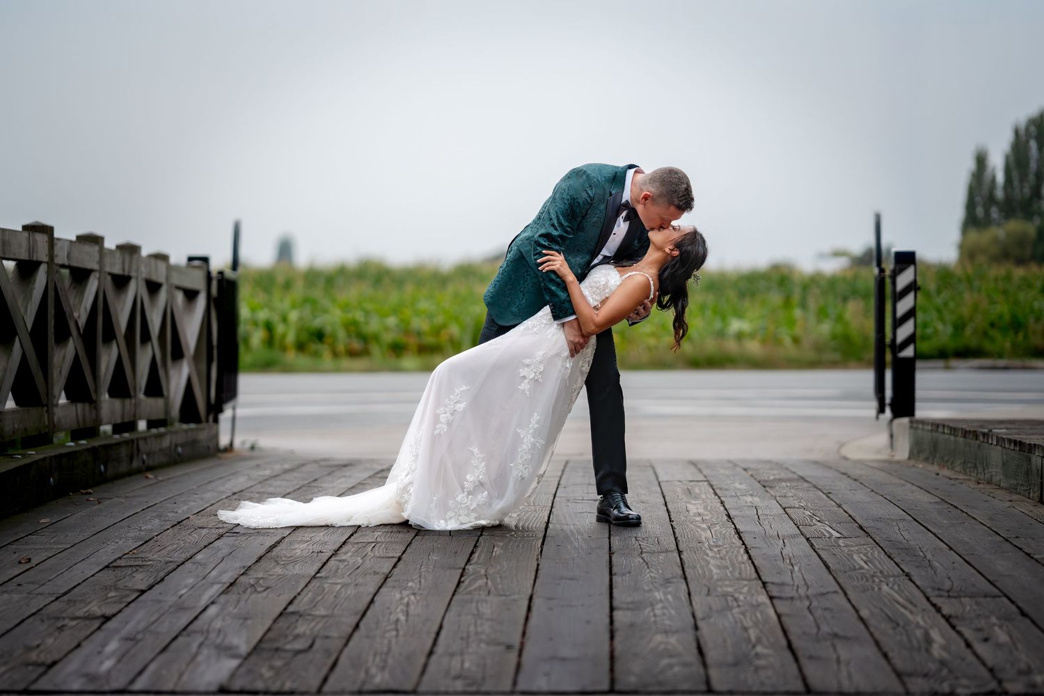 Groom dipping bride in romantic kiss on wooden dock in rain.