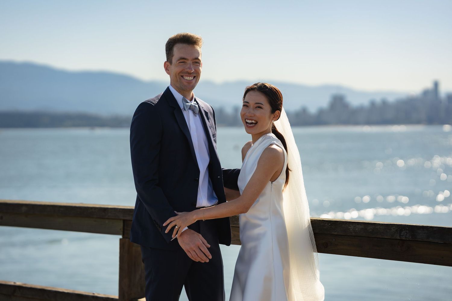 Happy couple in wedding attire smiling on a waterfront pier with mountain views and city skyline in background.