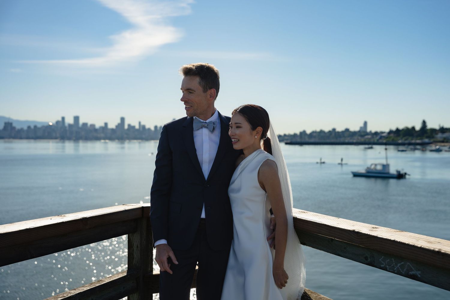 Bride and groom pose on a waterfront deck with a city skyline and blue sky in the background.