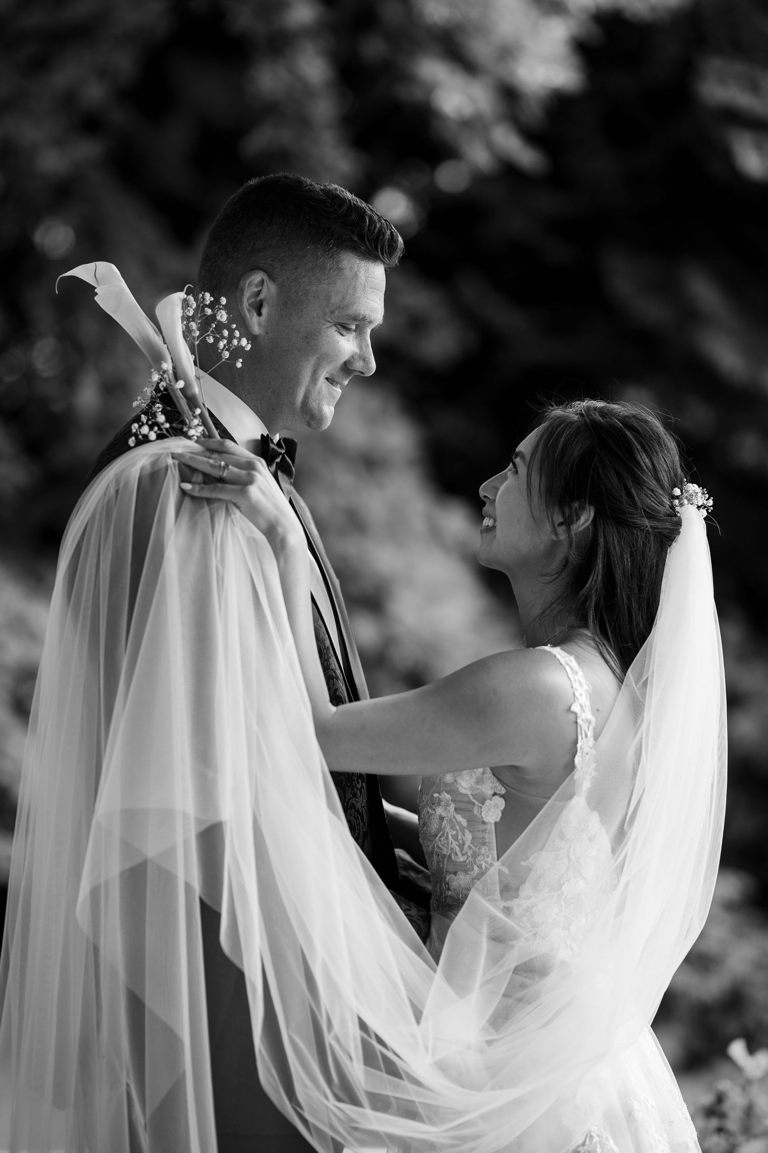 Black and white photo of a smiling bride and groom sharing a romantic moment outdoors, veil flowing around them.
