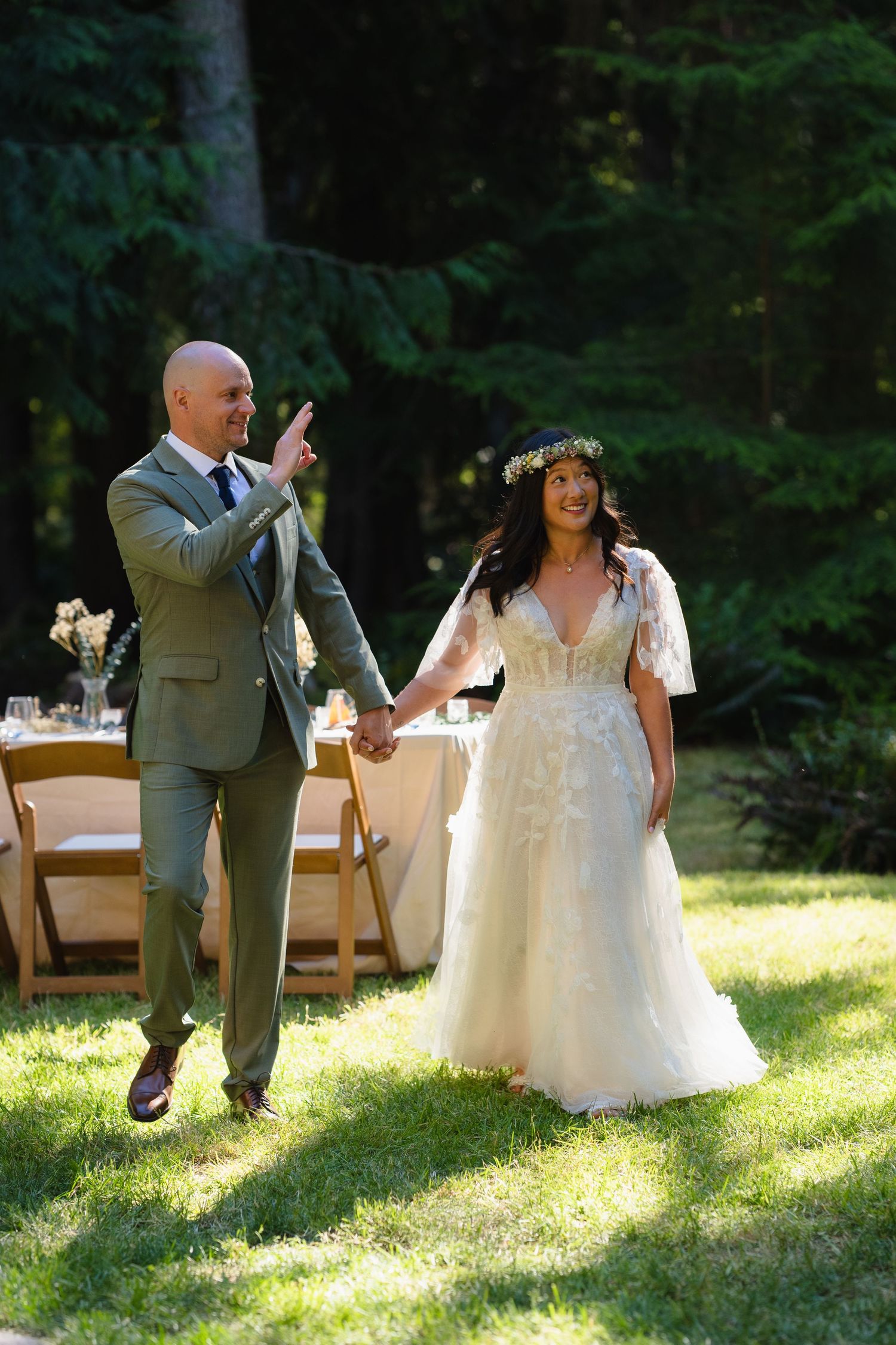 A smiling bride in a floral gown and groom in gray suit walk hand-in-hand at an outdoor wedding reception.