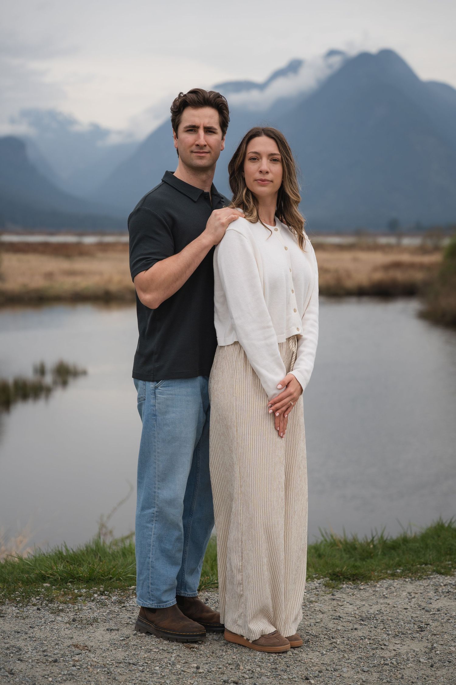 Couple posing together outdoors near a mountain lake, man in dark polo and jeans, woman in white outfit.