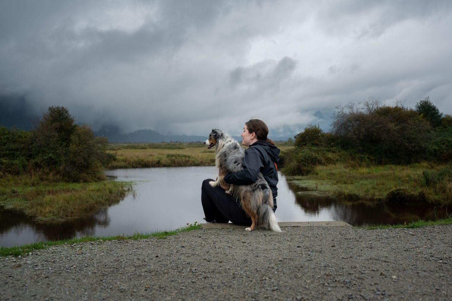 Woman sitting with her Australian Shepherd dog beside a calm pond under a dramatic cloudy sky.