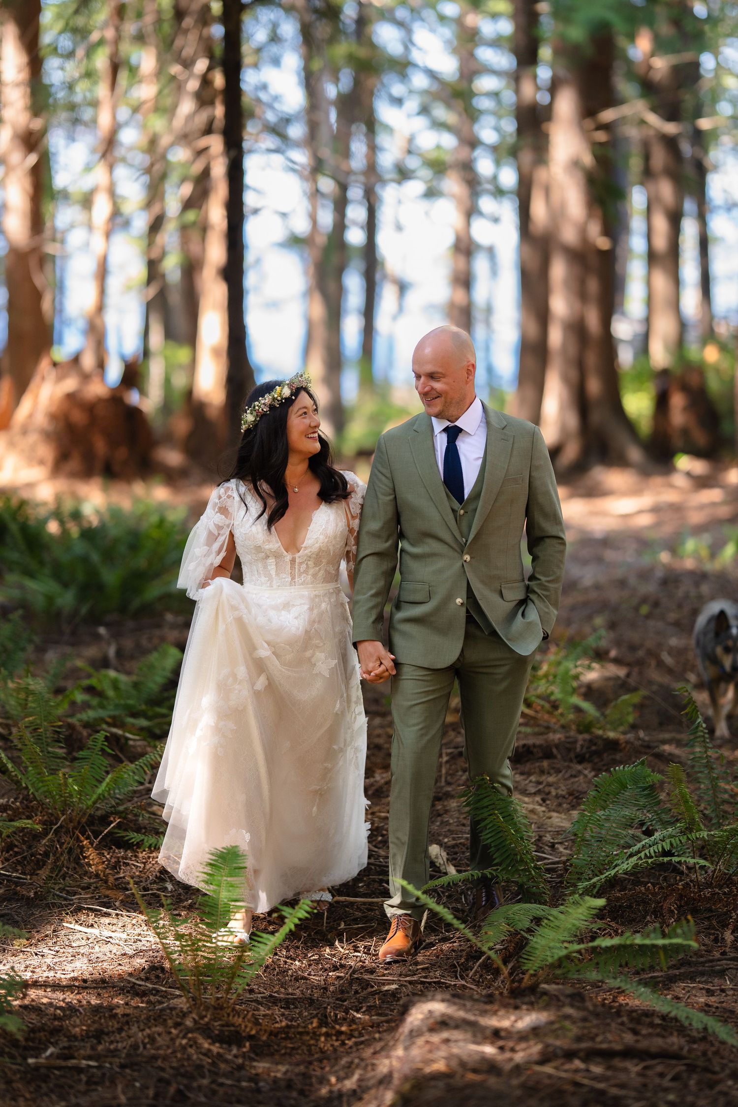 A bride and groom walk hand-in-hand through a sunlit forest, she in a lace gown and he in a sage green suit.