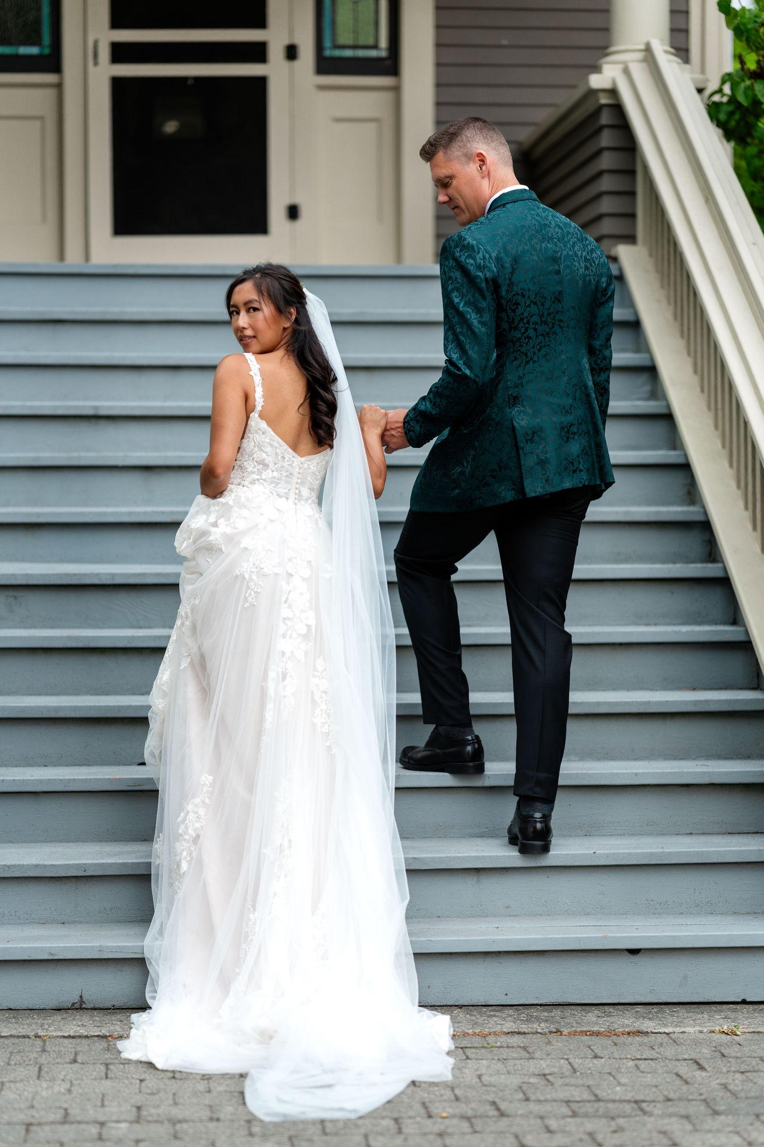 Bride in white lace gown and veil holds groom's hand in teal floral blazer as they walk up gray stairs.