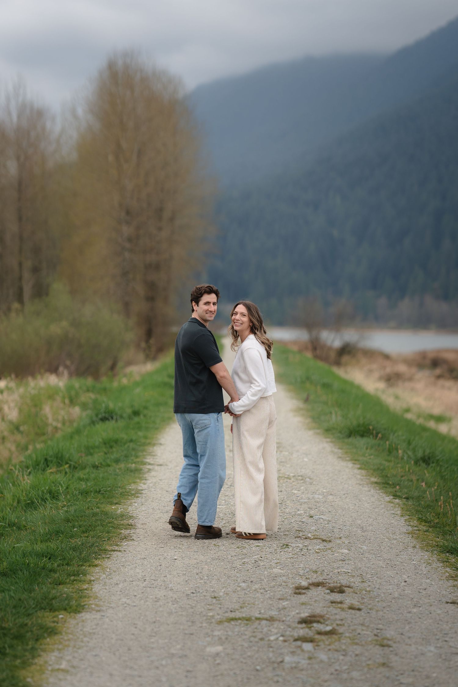 Couple holding hands on a scenic gravel path with mountains and lake in background.
