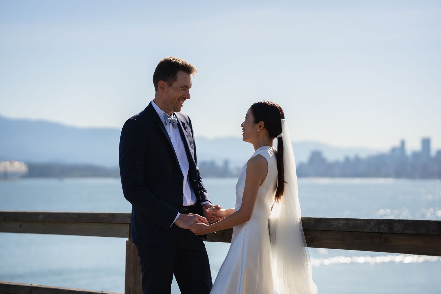 Bride and groom hold hands on waterfront pier with city skyline and mountains in background on wedding day.