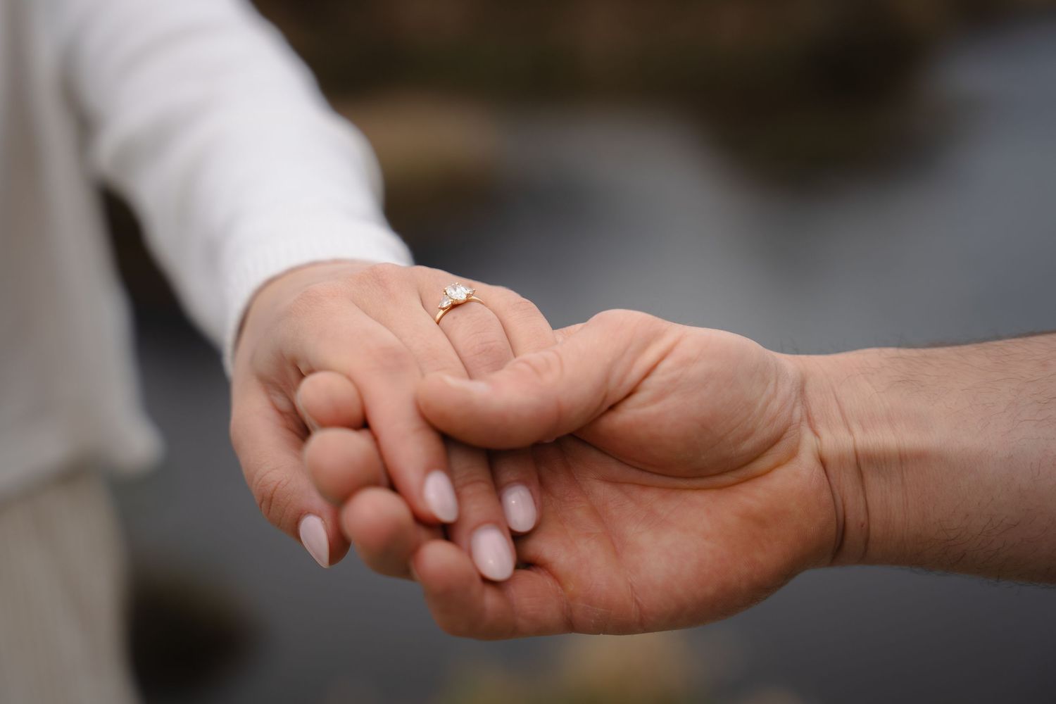 Close-up of two people holding hands, one wearing a gold engagement ring, with a blurred natural background.