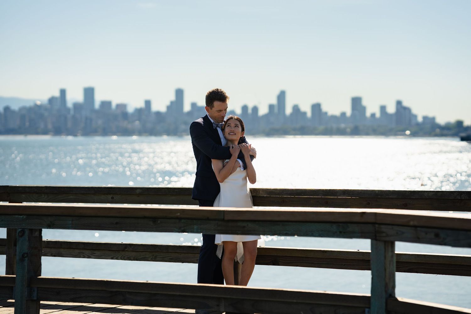 Couple embracing on a wooden pier with a stunning city skyline and sparkling water in the background.
