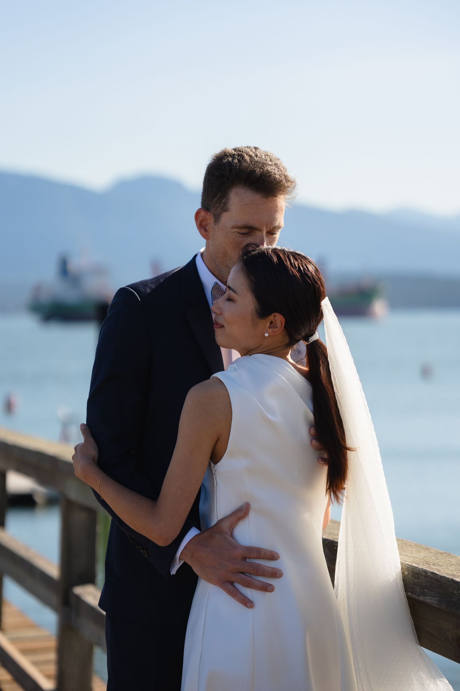 Bride and groom share a romantic embrace on a waterfront pier with mountains and a ship in the background.