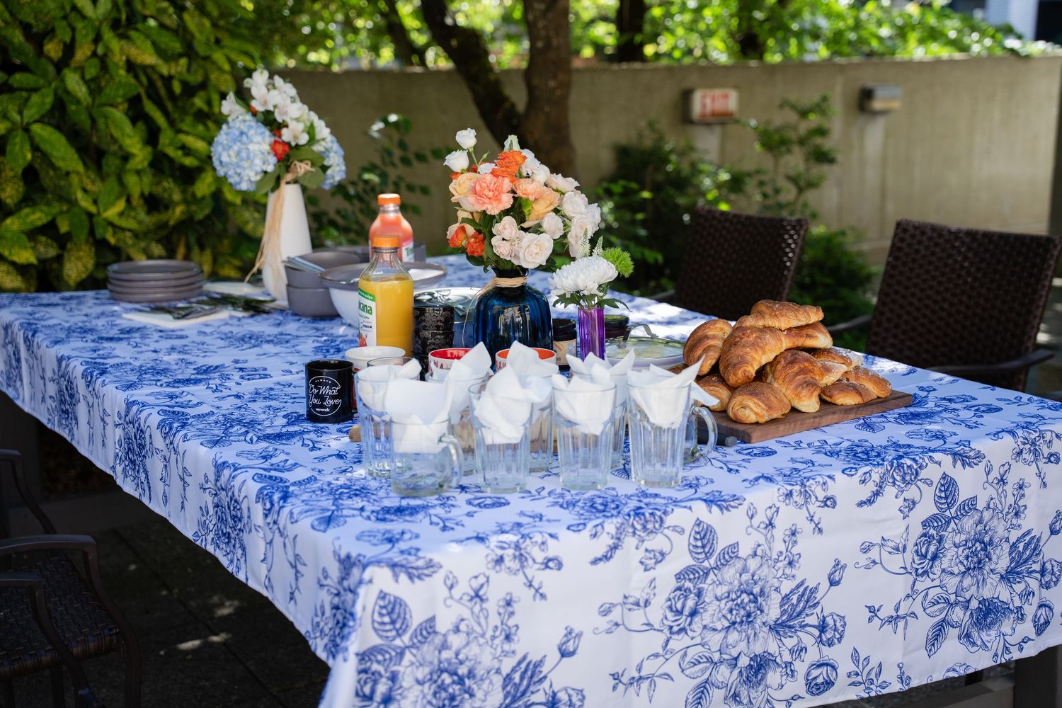 Outdoor breakfast table with blue floral tablecloth, croissants, fresh flowers, juice, and glassware set in a garden setting.