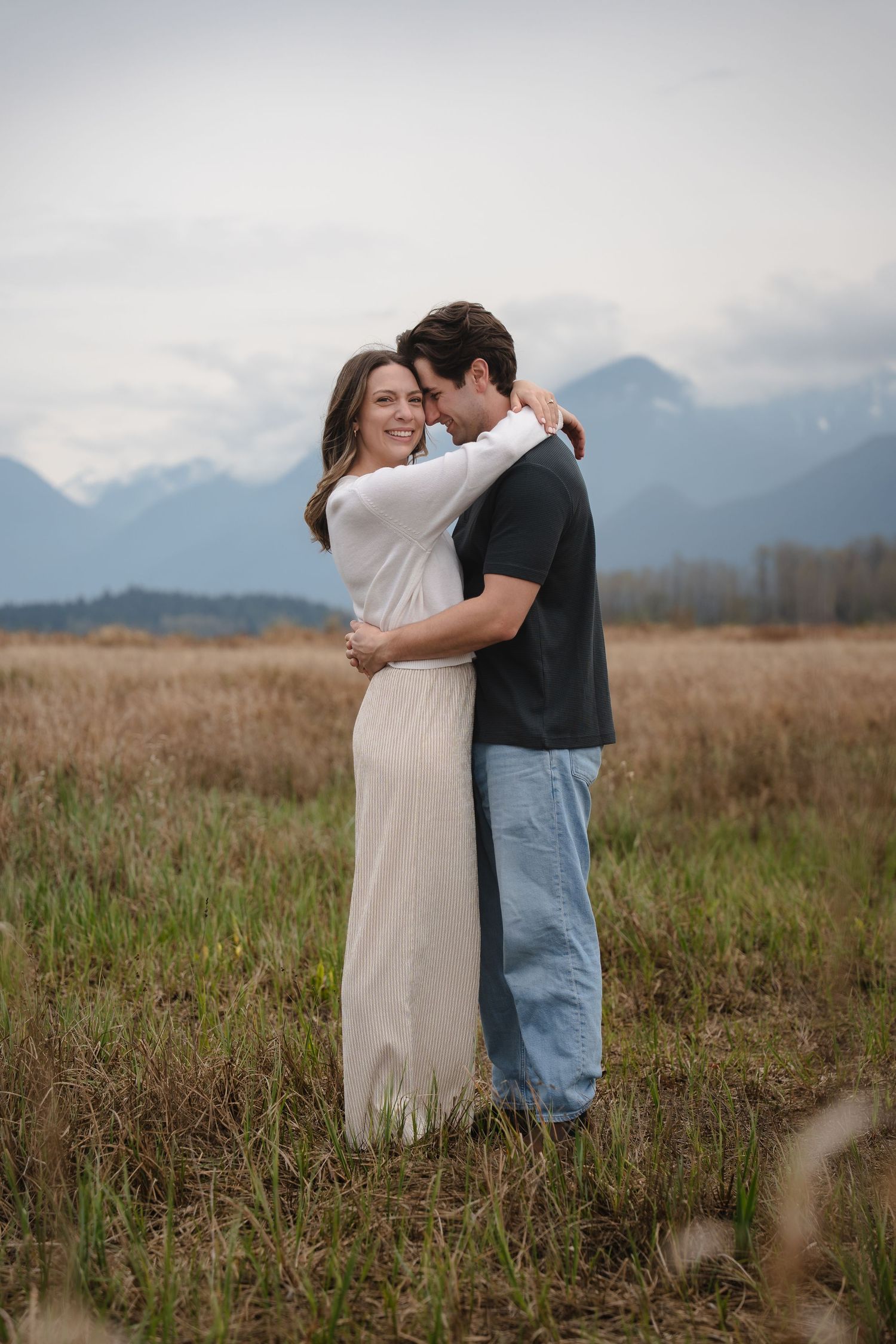 Happy couple embracing in golden field with stunning mountain backdrop during romantic outdoor engagement photo session.