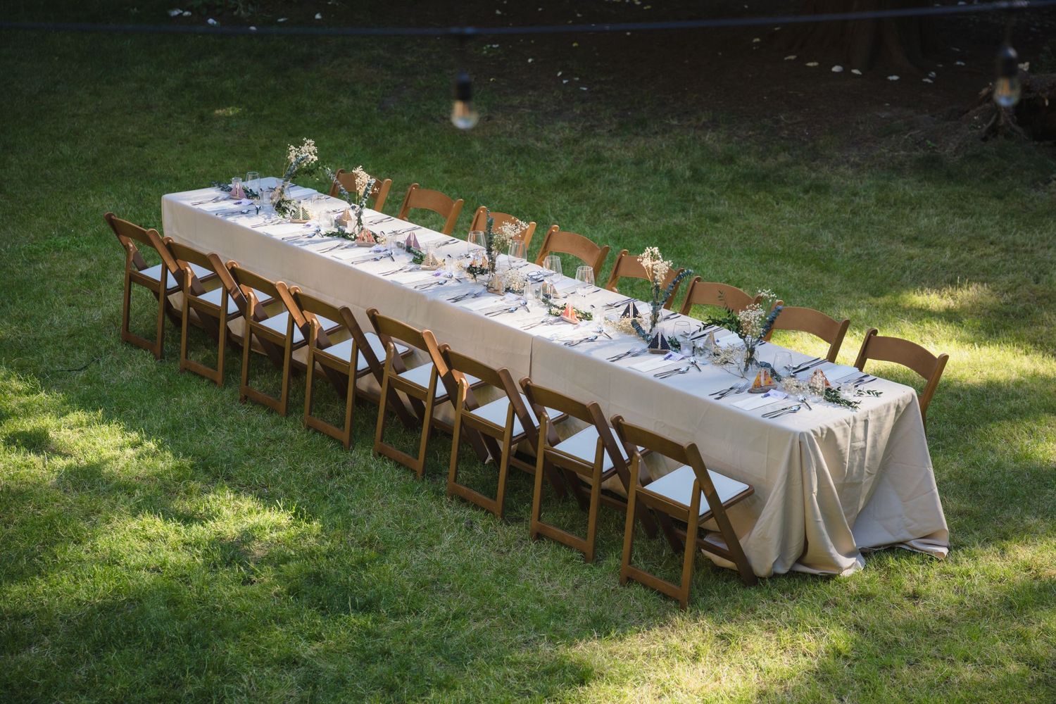 Elegant outdoor wedding reception table set on grass with wooden folding chairs and floral centerpieces.