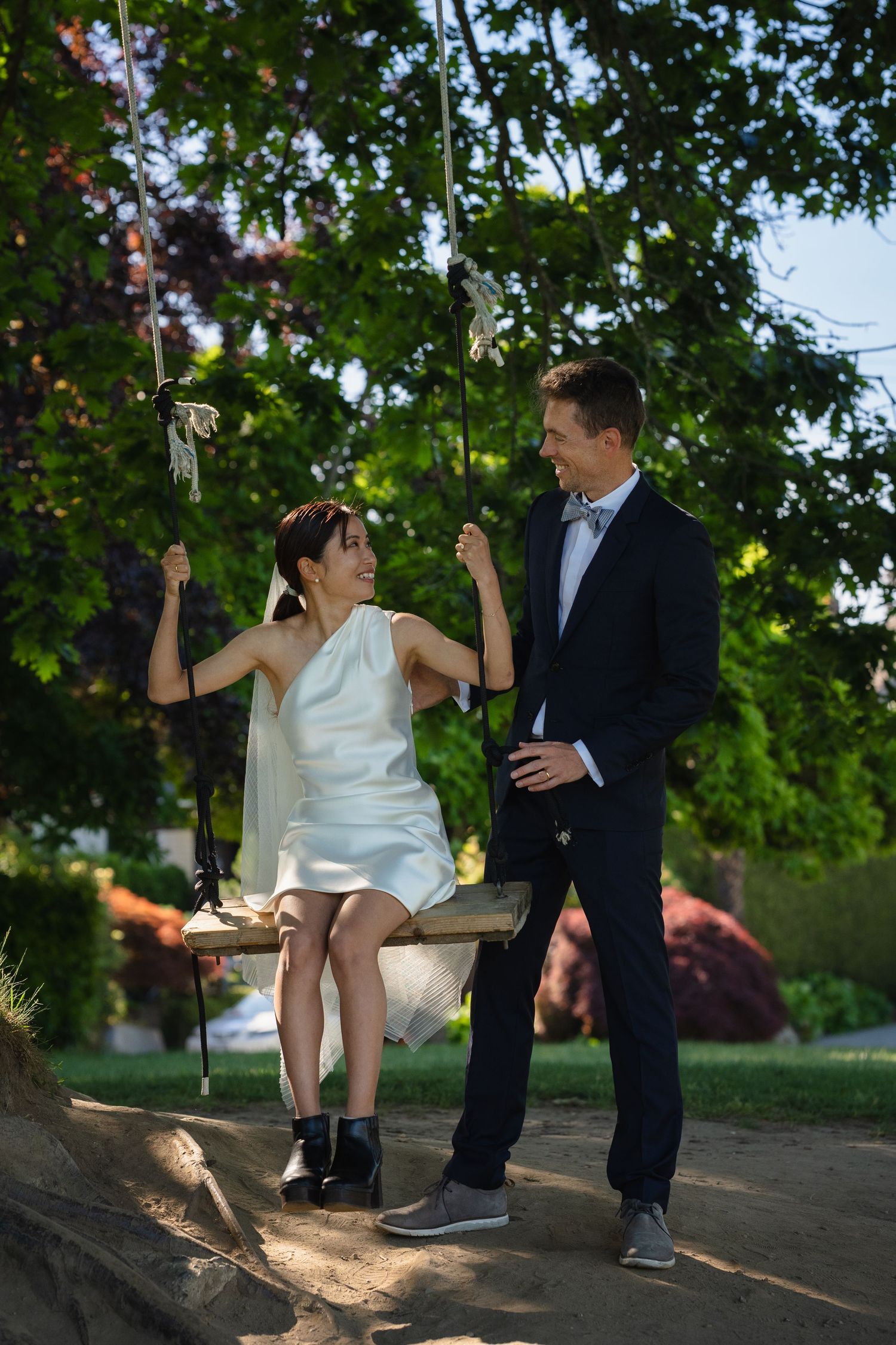 Bride in white dress sits on tree swing while groom in navy suit stands beside her in lush garden.