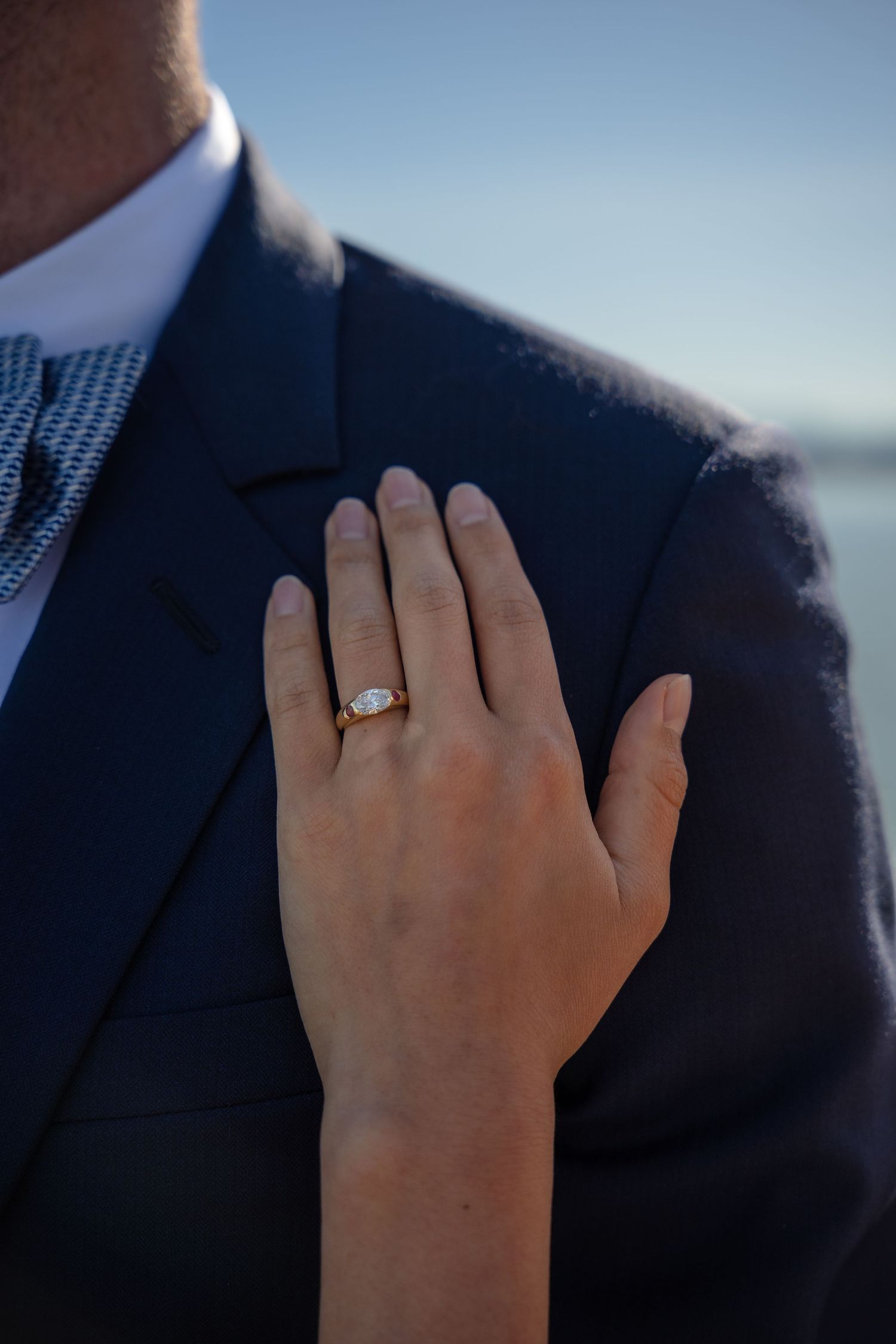 Close-up of a woman's hand displaying an oval diamond engagement ring resting on a man's navy blue suit jacket.