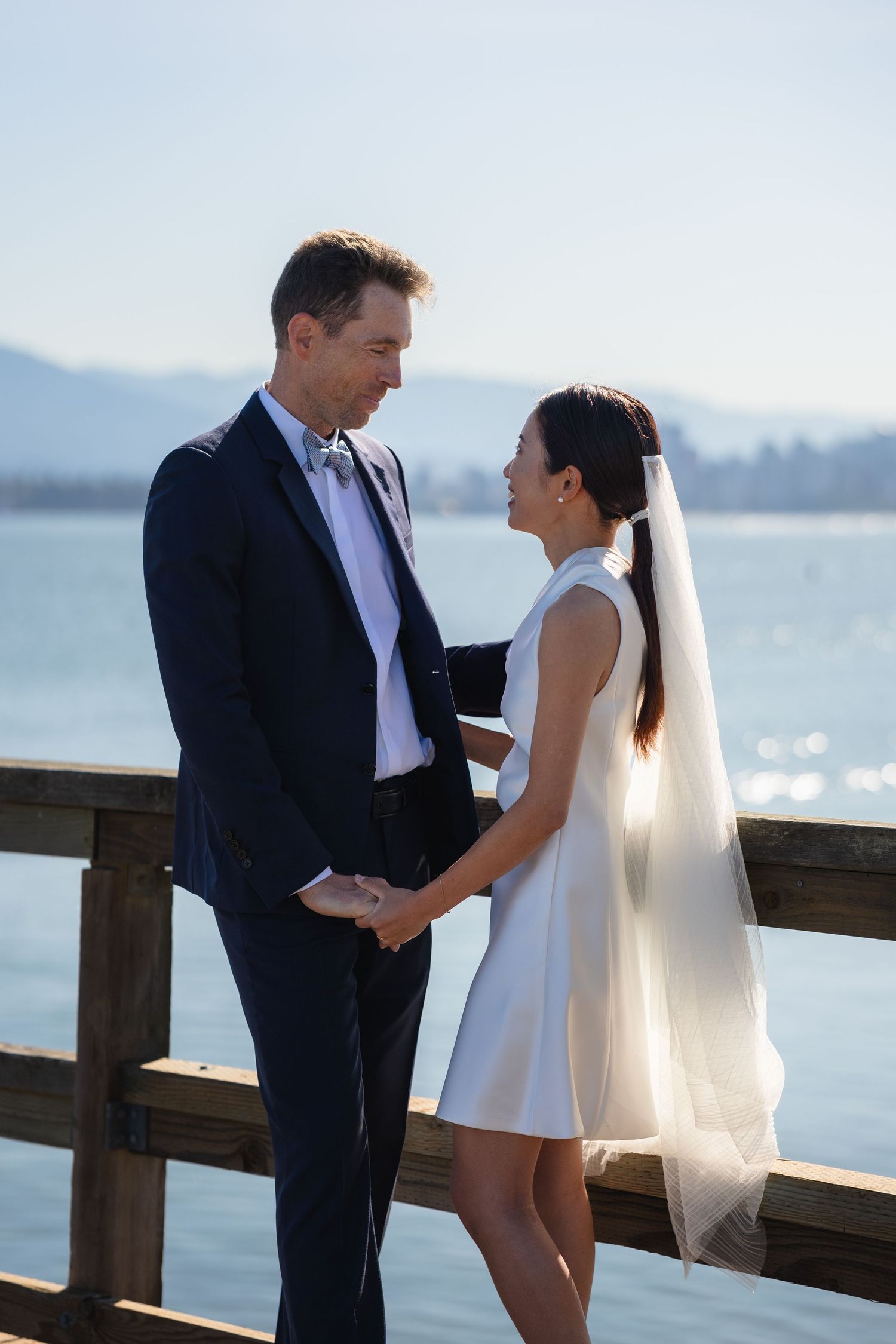 Bride and groom holding hands on a waterfront pier, exchanging vows in wedding attire with mountains in the background.