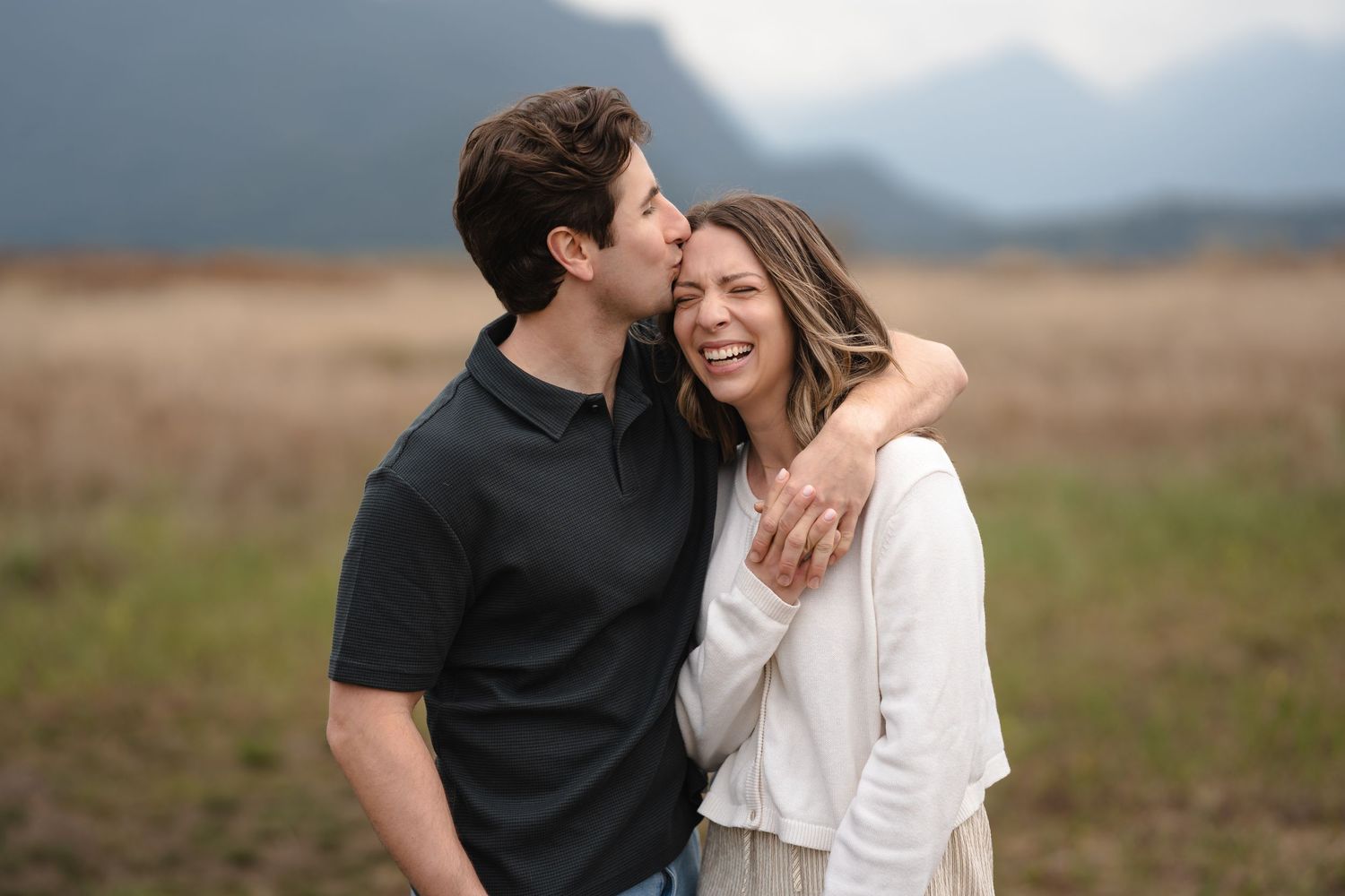 A couple shares a romantic moment in a scenic field, man kissing woman's cheek as mountains loom in the background.