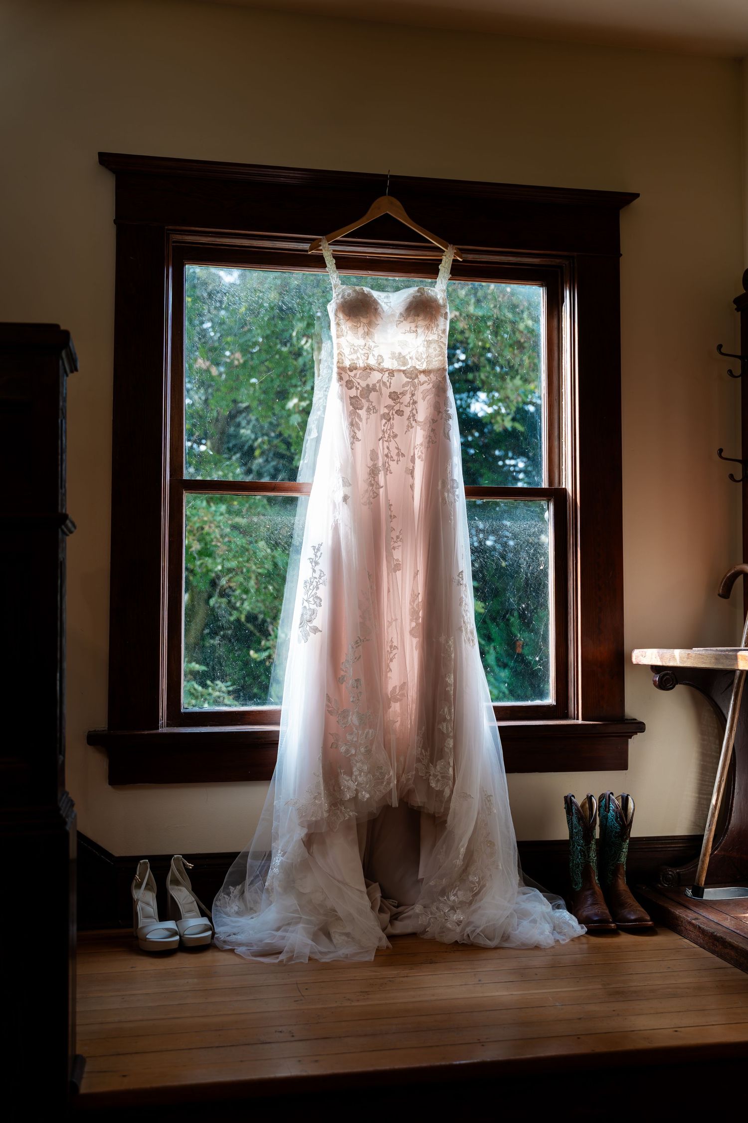 Elegant lace wedding dress hanging on window hanger with bridal shoes below.
