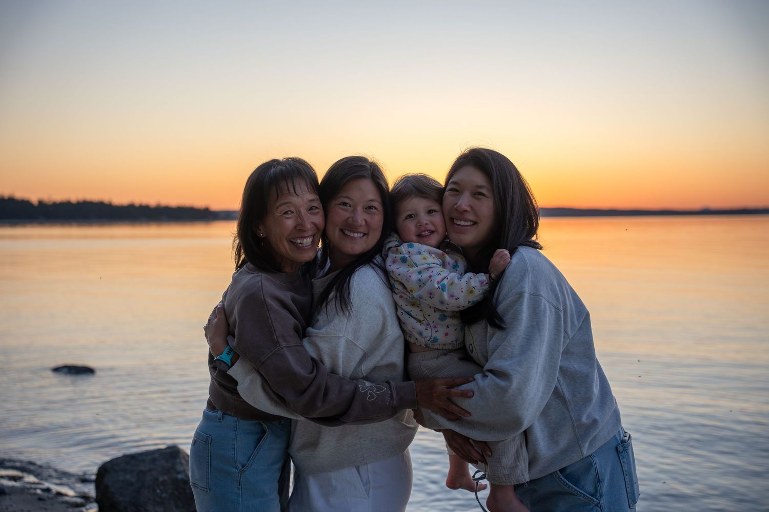 Four women smile and embrace during a golden sunset by the water in a heartwarming group photo.