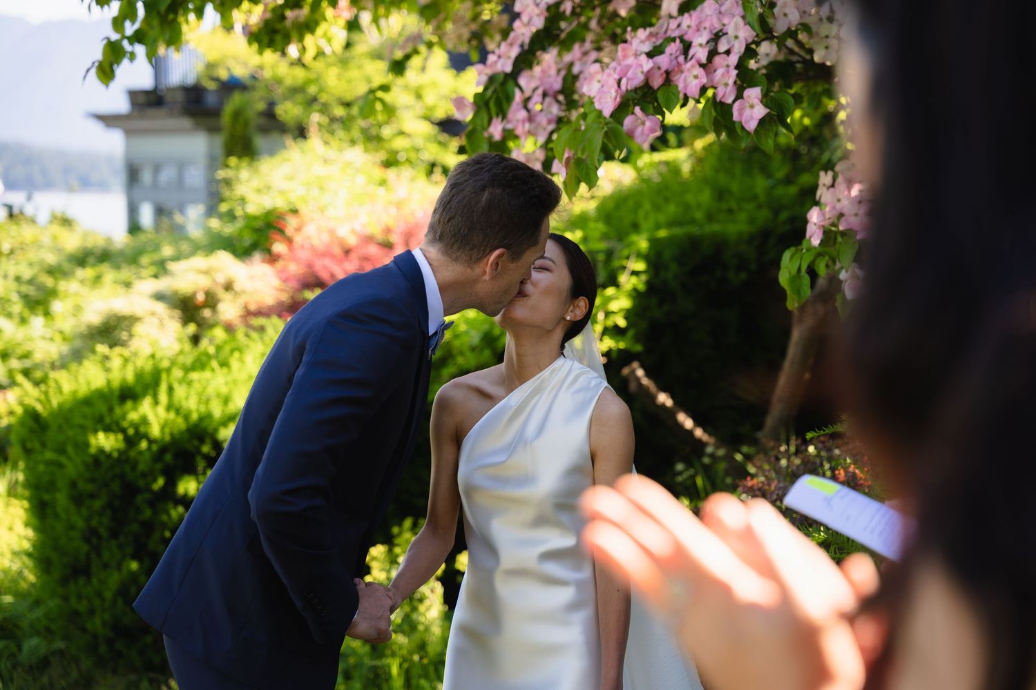 Couple sharing a kiss at outdoor wedding ceremony surrounded by lush greenery and pink flowering trees.