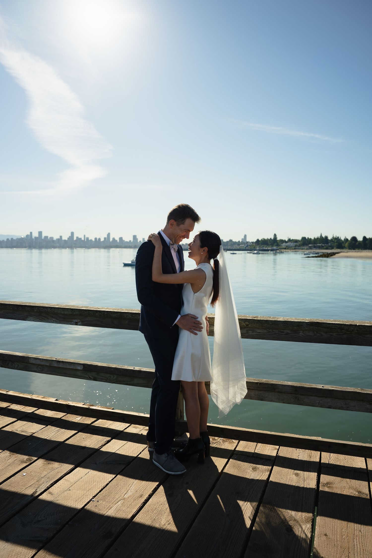 Bride and groom embrace on a waterfront pier with a city skyline and blue sky in the background.
