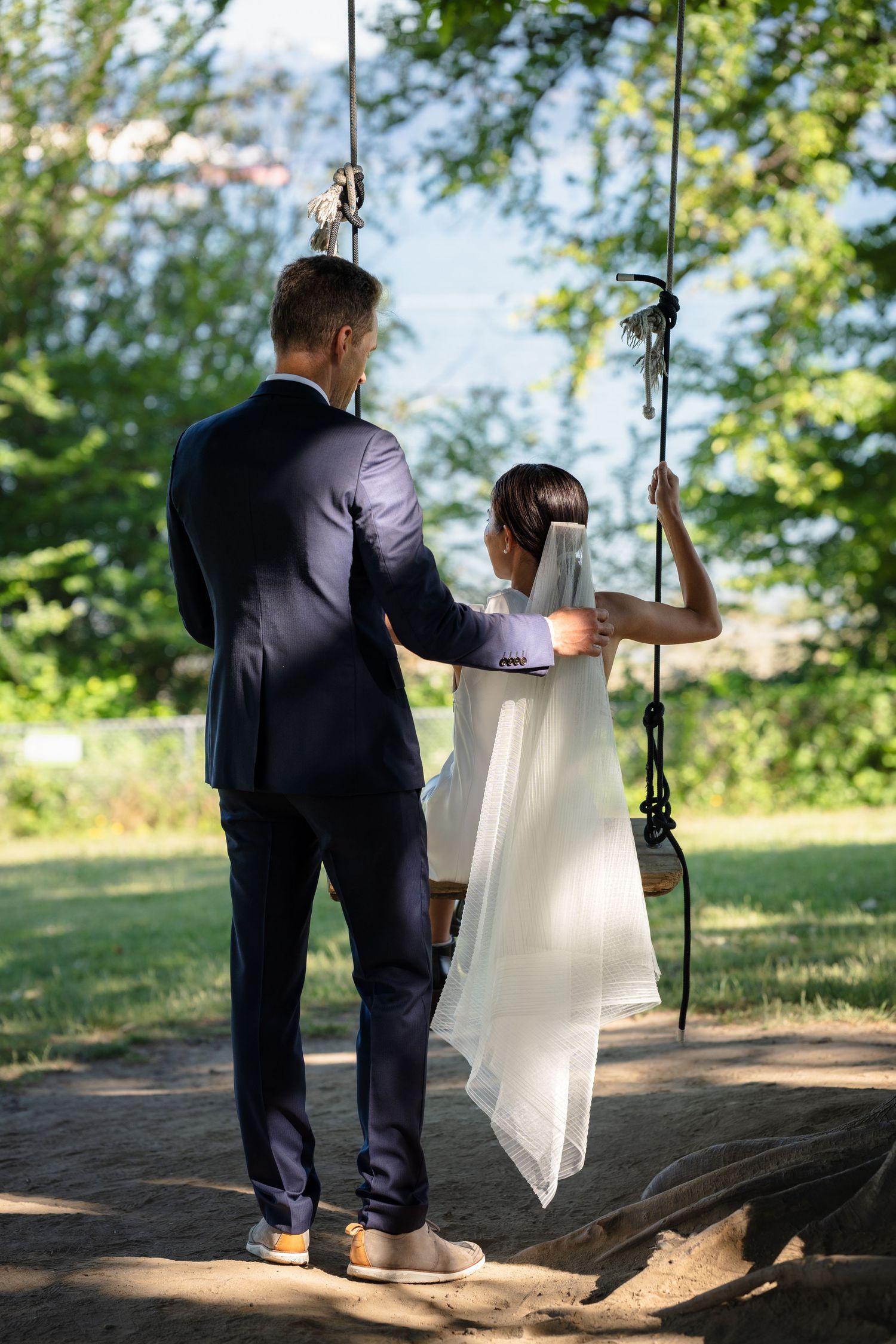 Bride in white dress sits on rope swing as groom in navy suit stands beside her in a sunny green park.