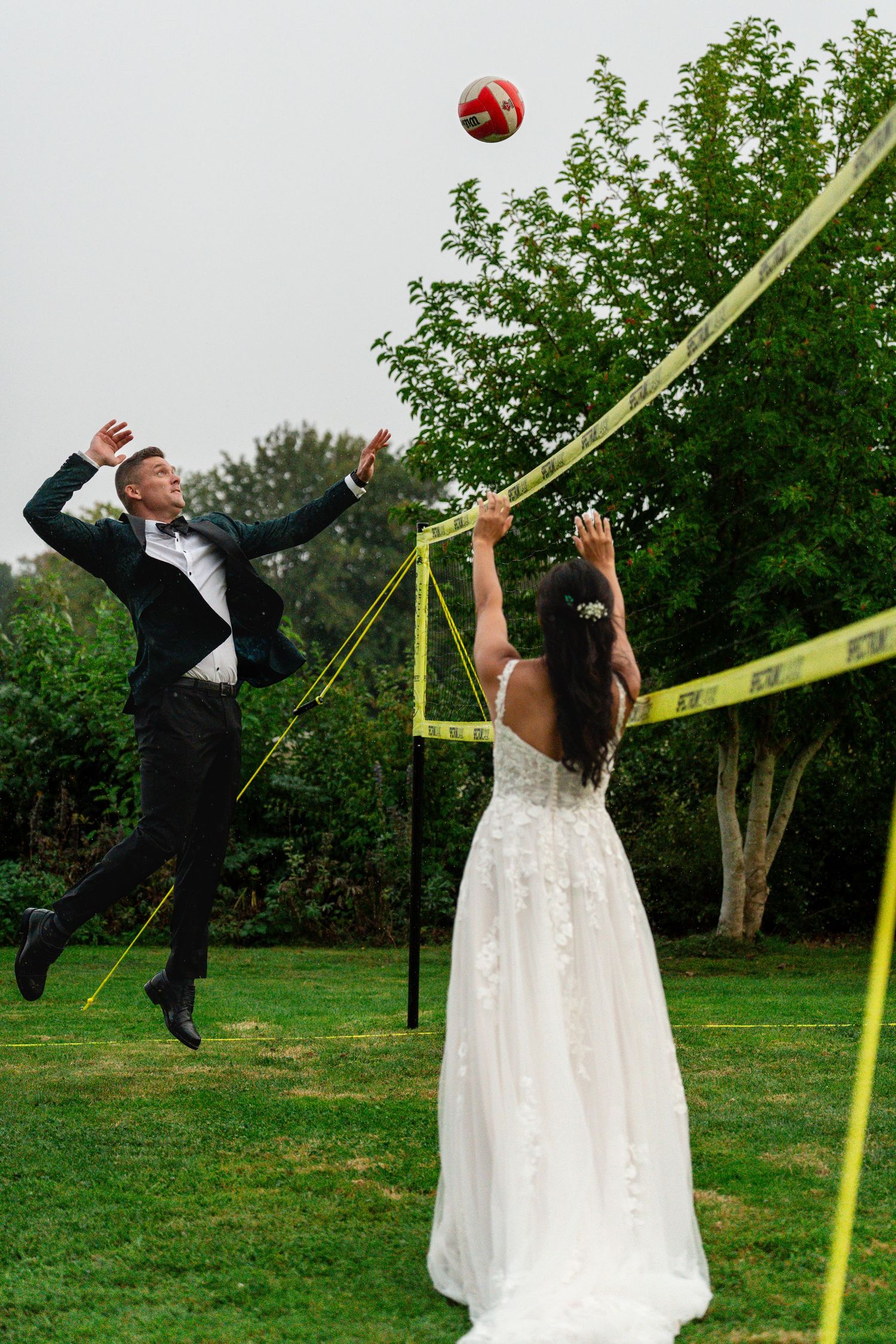 Bride and groom playing volleyball at wedding reception, groom jumping to spike red ball over yellow net.