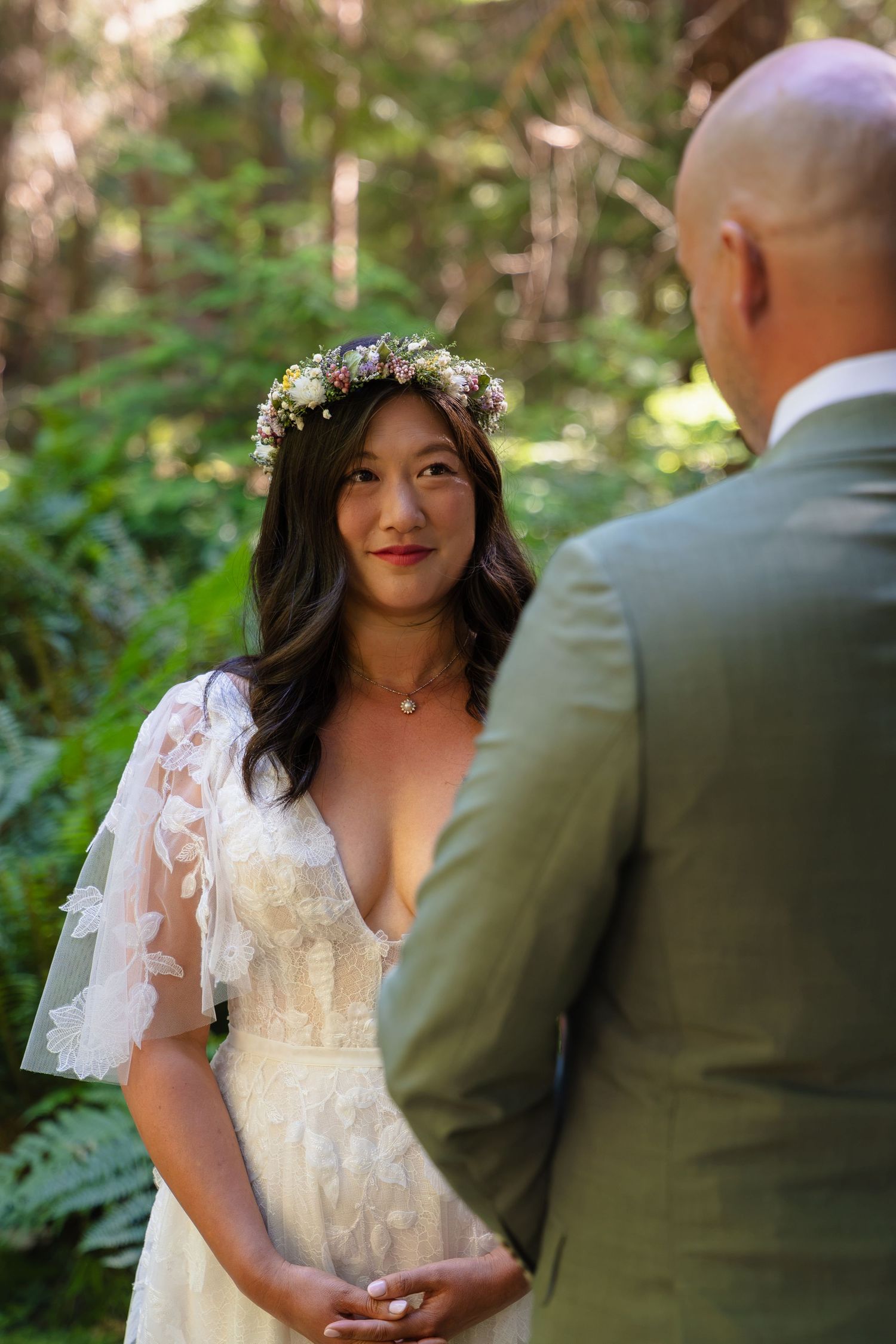 Bride in lace dress and floral crown smiles at groom during outdoor forest wedding ceremony.