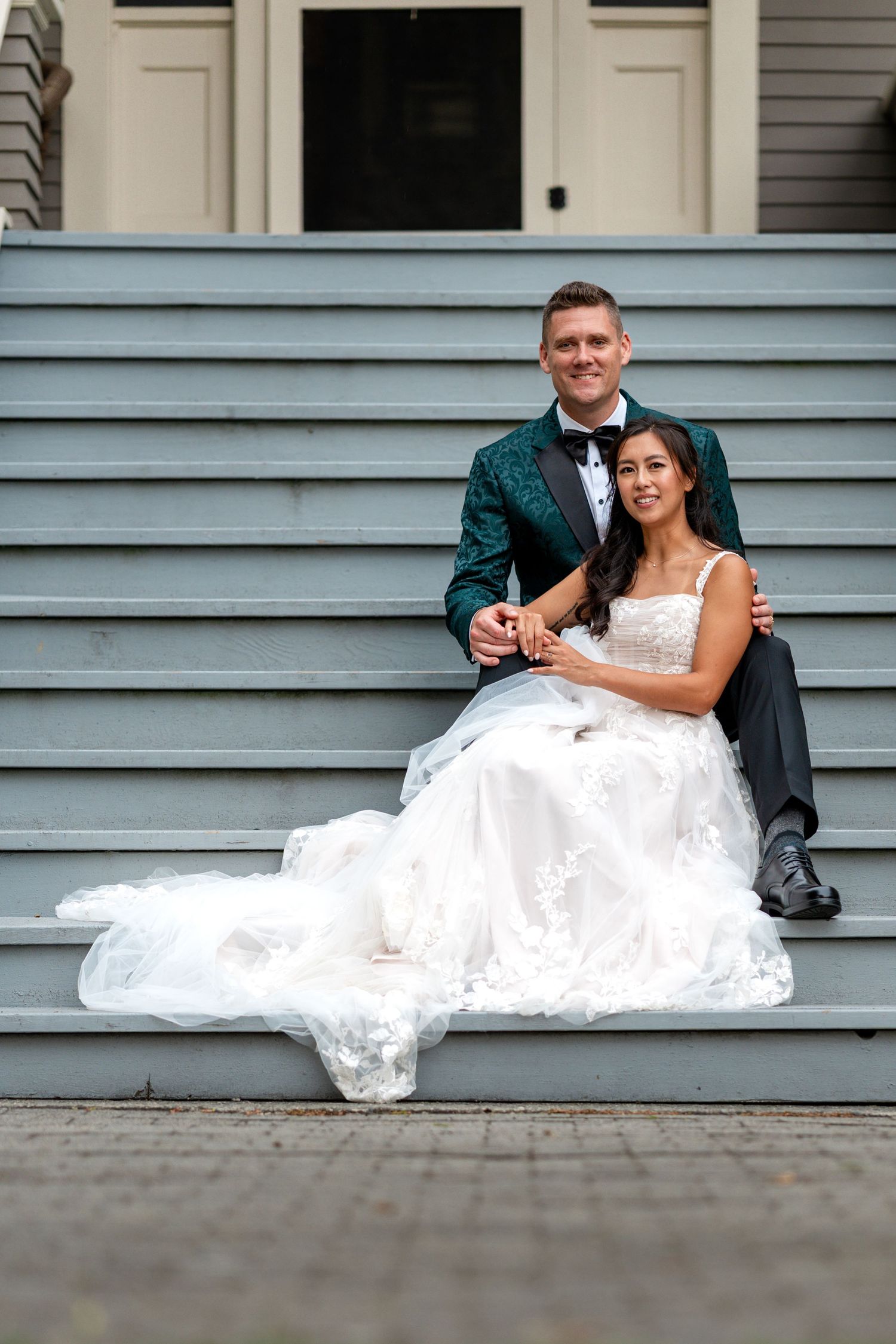 Bride and groom sitting together on grey steps with brides white gown spread elegantly around them.