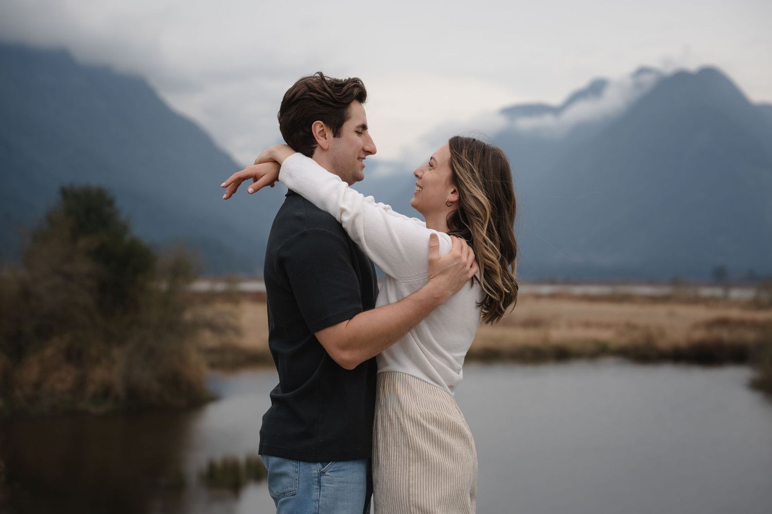 Couple embracing and smiling near a mountain lake with misty peaks in the background.