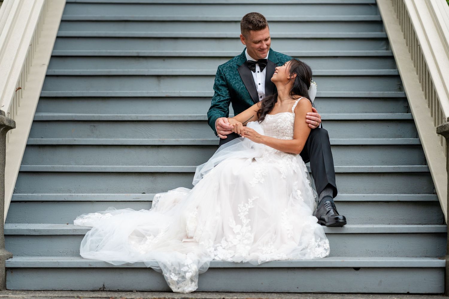 Bride and groom sitting together on gray outdoor steps, wearing wedding attire, sharing a romantic moment.