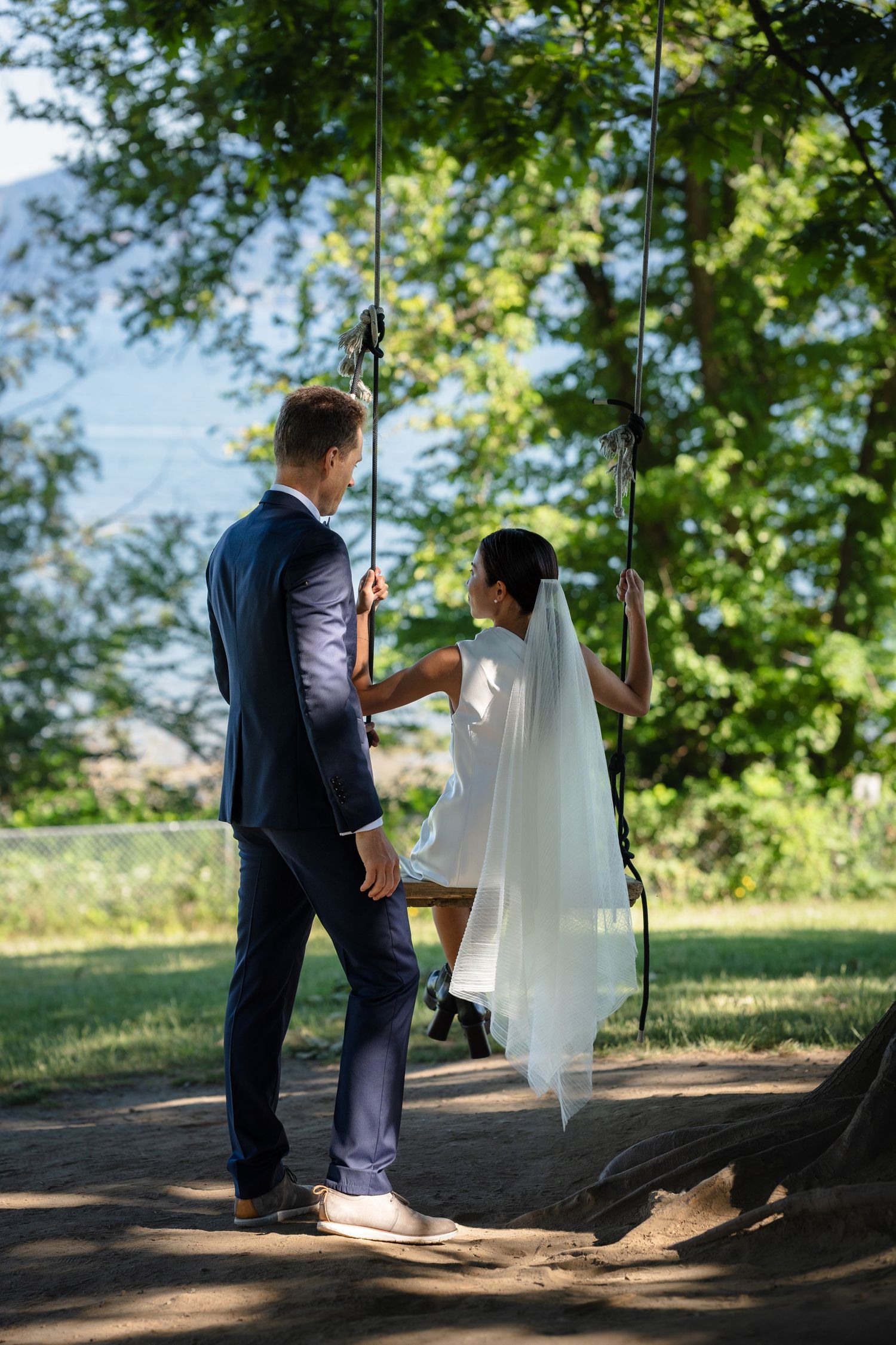 Bride in white dress sitting on a swing as groom in navy suit stands beside her in a lush green park.