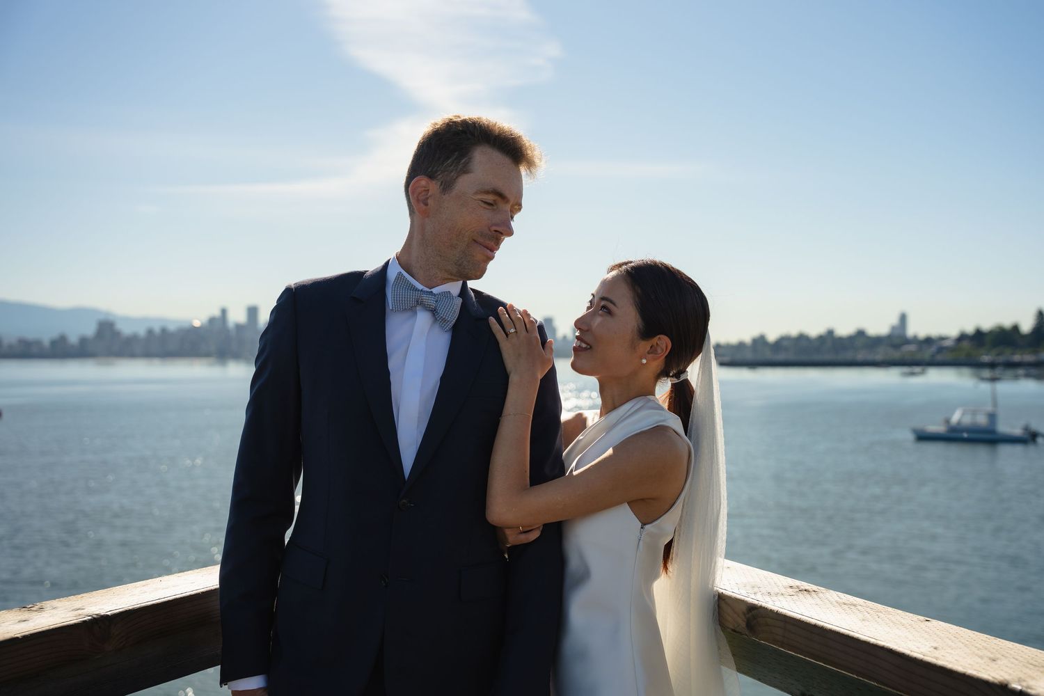 Bride and groom share a romantic moment on a waterfront deck with a blue sky and city skyline backdrop.