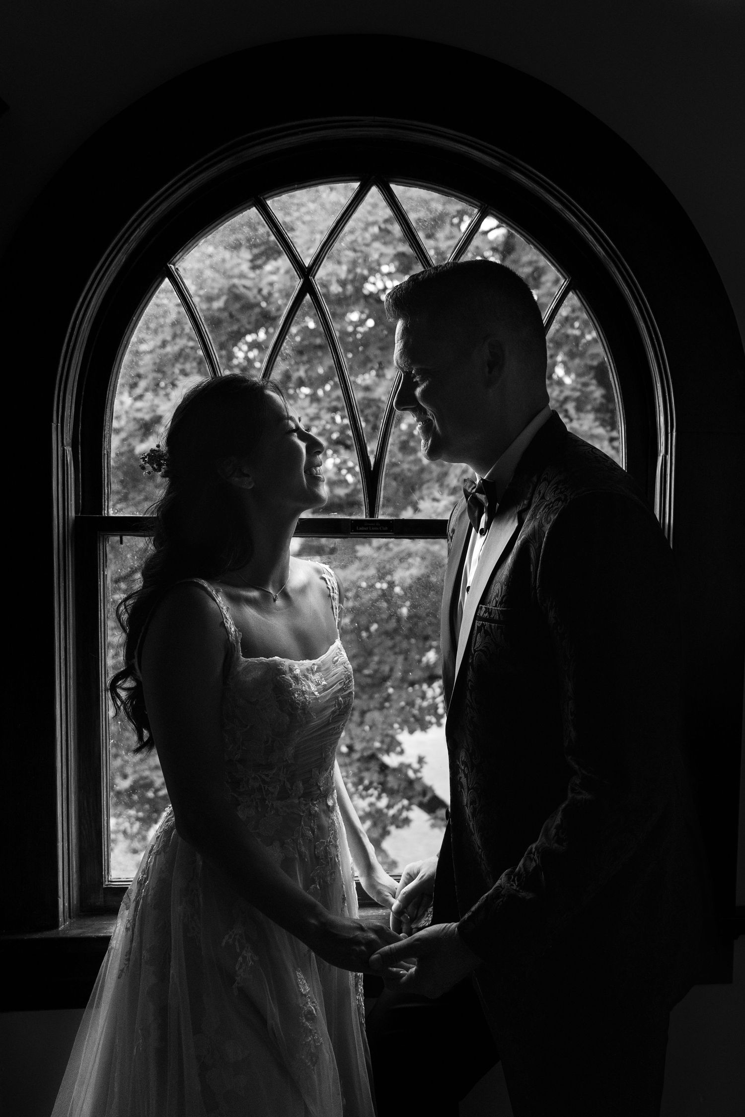 Romantic black and white silhouette of bride and groom holding hands before an arched church window.