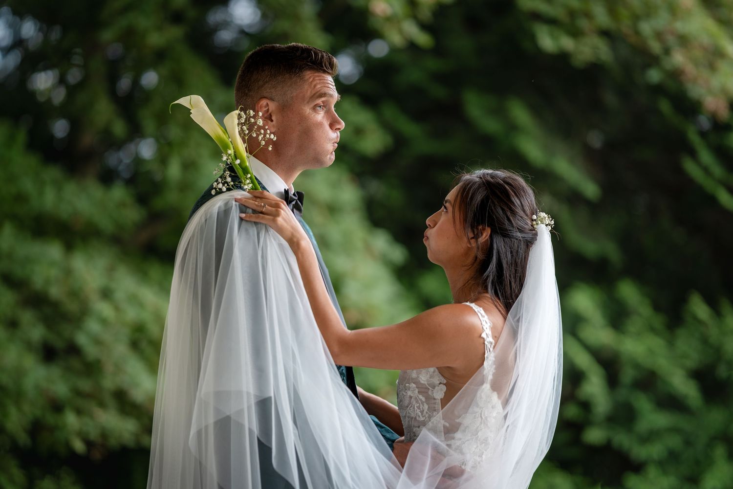 Bride adjusting grooms boutonniere while holding calla lily bouquet under her veil in outdoor setting.