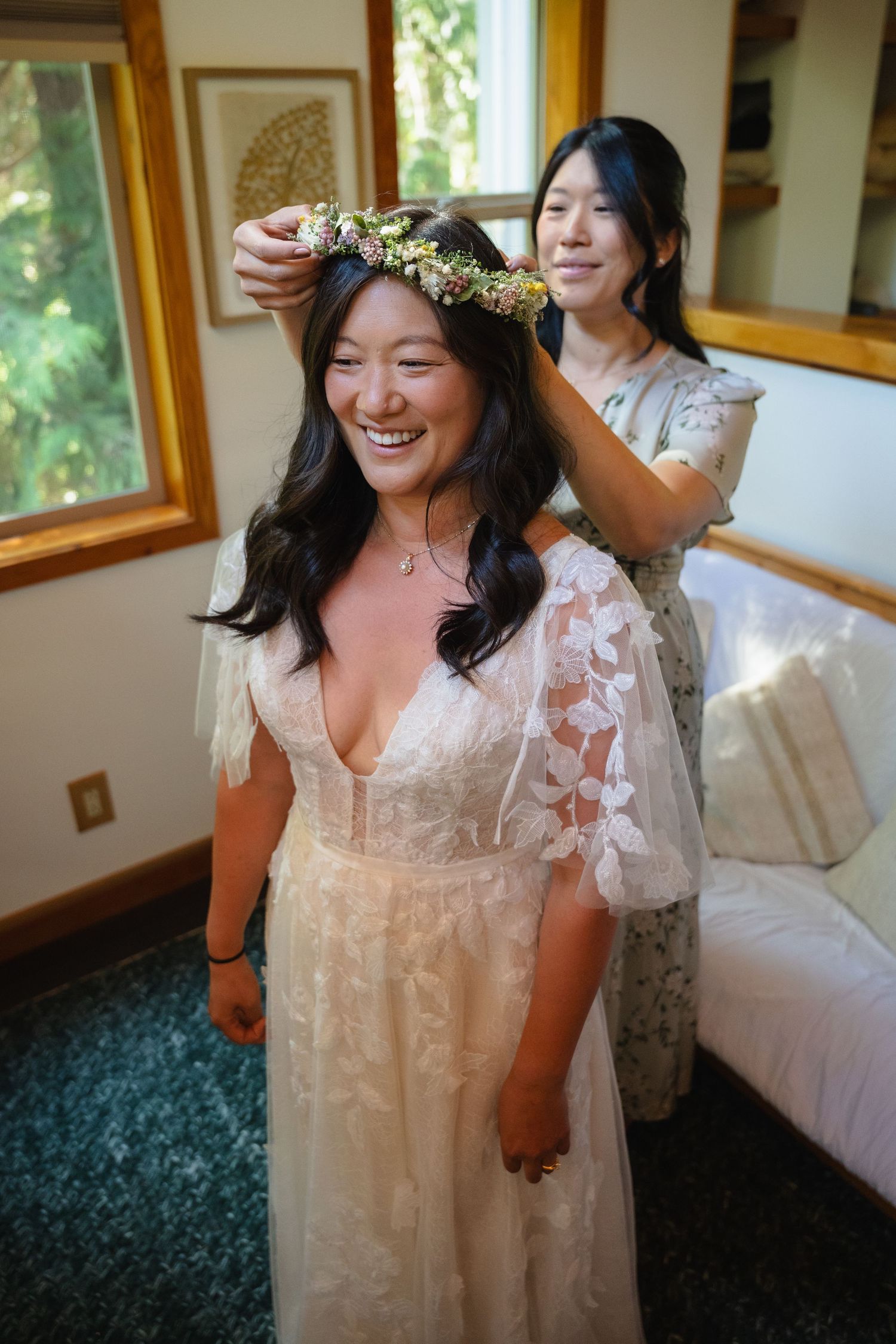 Smiling bride in lace wedding dress gets flower crown placed on her head by a woman in a floral dress indoors.