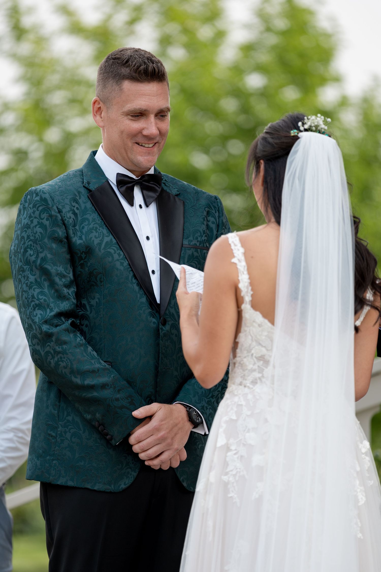 Groom in green floral tuxedo smiling at bride in white lace gown and veil during outdoor wedding ceremony.