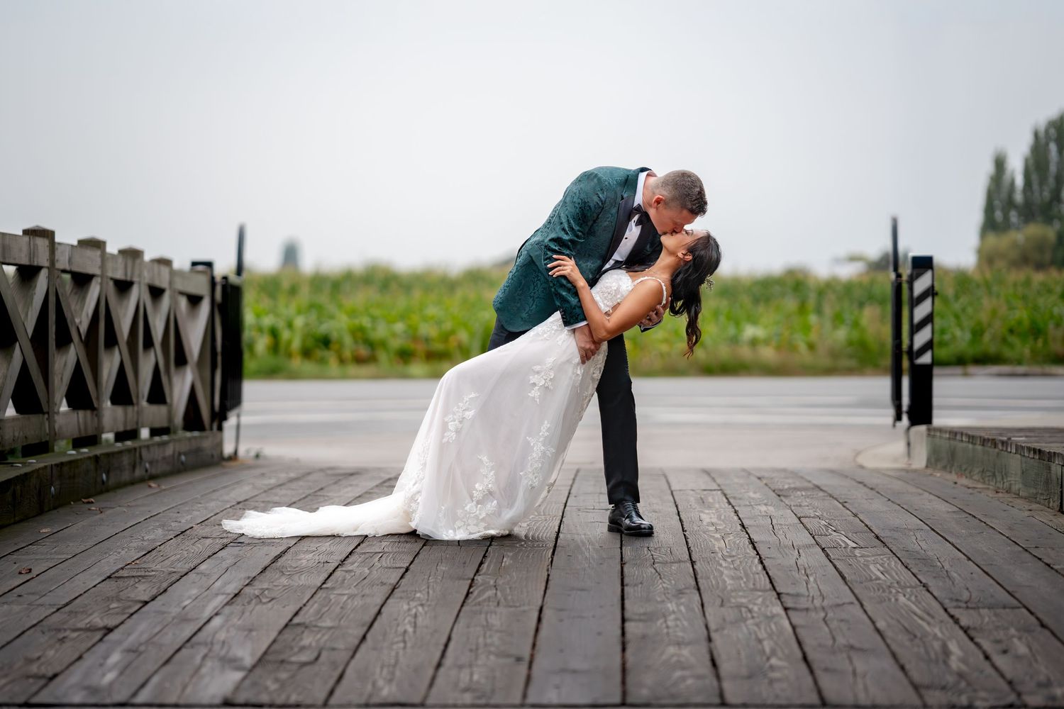 Bride and groom share a romantic dip kiss on a wooden dock, with the groom in a teal suit and bride in a white gown.